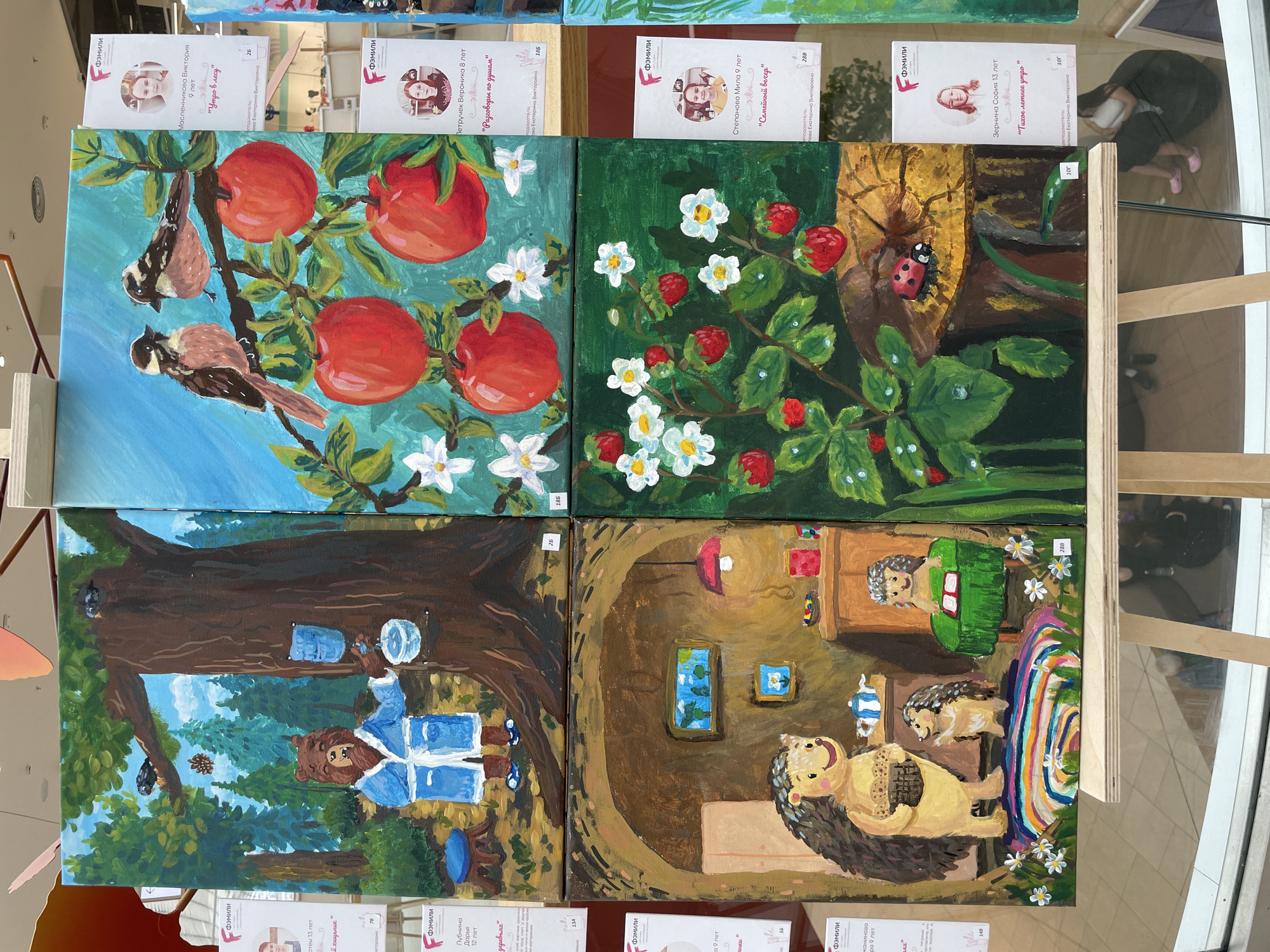 The image displays an exhibition of four paintings, each depicting a whimsical scene, arranged in a 2x2 grid on a wooden easel. The exhibition appears to be held in a public indoor space, possibly a mall or gallery, indicated by the glass reflections of people and the tiled floor in the background. A sign in the upper left corner features the logo "ФЭМИЛИ" (FEMILY) and text in Cyrillic, suggesting the exhibition is related to a children's art group or competition.
The top-left painting shows a brown bear wearing a blue bathrobe, holding a cup and pouring water from a dispenser attached to a large tree. Two birds are perched on a branch above. This painting is labeled with a number "26".
The top-right painting features two small birds on a branch laden with ripe red apples. White flowers are also visible. This painting is labeled with the number "186".
The bottom-left painting depicts a cozy interior scene with two anthropomorphic hedgehogs. The larger hedgehog, carrying a basket, stands on a colorful rug, while a smaller hedgehog sits at a table with a book. A lamp is on the wall, and a window shows a blue sky with trees. This painting is labeled "288".
The bottom-right painting showcases a vibrant strawberry plant with red strawberries and white flowers, some with water droplets on their leaves. A ladybug is prominently featured on a piece of wood. This painting is labeled "10r".
Beside each painting, there are information cards with a portrait of a child, their name, age, and the title of their artwork in Russian. The text on the cards also includes the name of an instructor. One card indicates "Ярцева Екатерина Викторовна" as the instructor for multiple artworks. Another card mentions "Степанова Мила 9 лет" with the title "Тихое летнее утро". The presence of the "ФЭМИЛИ" logo, the Russian text on the cards, and the names of the children suggest a Russian origin for the exhibition. While the specific location within the city of Perm, Russia is not explicitly stated, the overall context points to a local event or exhibition in that city. The lighting is bright, suggesting an indoor environment with artificial lighting. Some reflections in the glass reveal people, including a person sitting on the floor in the background, and the tiled floor.