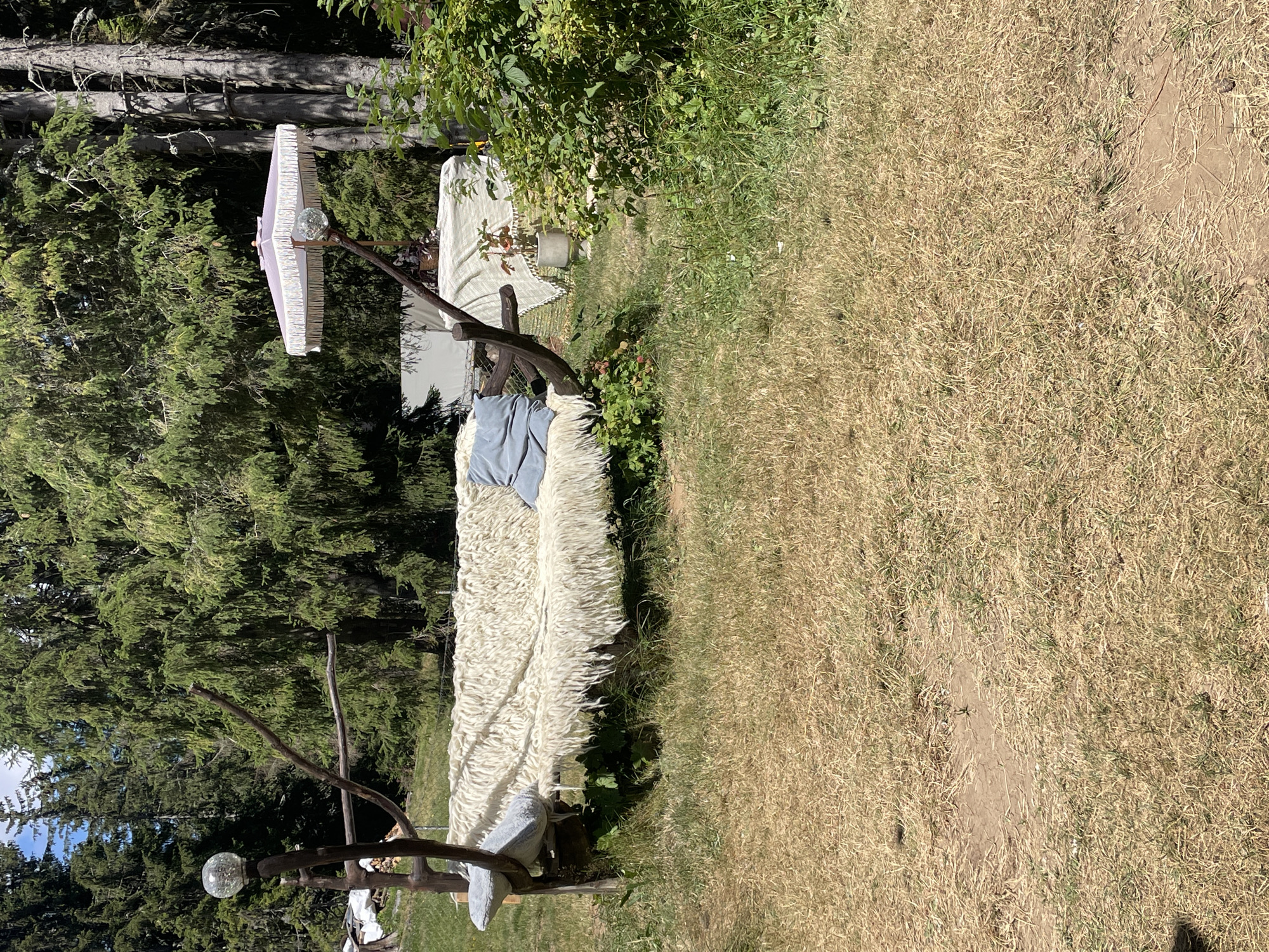 This image depicts a rustic outdoor seating area, likely in a natural setting such as a forest or meadow, with a prominent wooden swing-style bench covered with a thick, shaggy off-white throw and two light blue pillows. To the right of the bench, a small table is set with a white tablecloth, and a potted plant with red flowers. An umbrella with white fringe stands behind the table. The environment is characterized by dry, golden grass in the foreground and lush green pine trees in the background, suggesting a sunny, warm day. There is no indication of the city of Smolyan, Bulgaria, in the image itself.