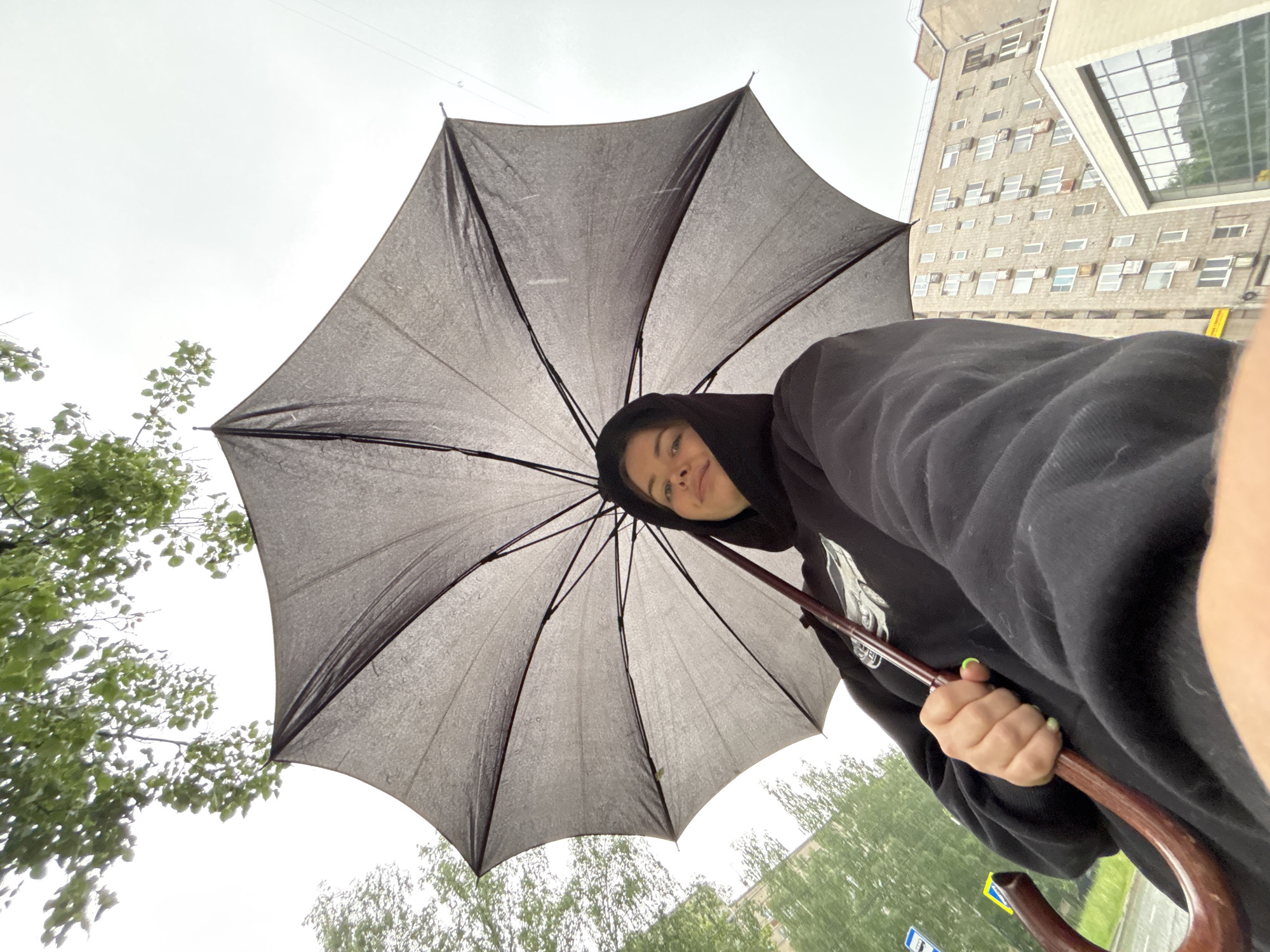 This low-angle, wide-shot image captures a person holding a large, open black umbrella, appearing to be taking a selfie or looking directly at the camera. The subject is a young individual, seen from the chest up, wearing a black hoodie with the hood pulled over their head. Their face is visible, featuring a slight, pleasant smile, and they have light green nail polish on their right hand, which is gripping the curved, brown handle of the umbrella. The umbrella itself is prominent, wet with what appears to be raindrops, indicating rain or recent rain.
The setting appears to be outdoors on an overcast day. The sky is bright but uniformly grey, suggesting a cloudy or rainy weather condition. To the left and bottom of the frame, there are green trees with fresh foliage. In the background, to the right, stands a tall, light-colored building with many rectangular windows of varying sizes and positions, giving it an older, possibly utilitarian or Brutalist architectural feel. A more modern glass structure is visible near the top right of this building. A blue and white road sign is partially visible in the bottom right corner, suggesting a street or public area. The overall impression is a daytime scene in Izhevsk, Russia, where the individual is comfortable despite the inclement weather, perhaps enjoying a walk in the rain. No specific text is clearly legible within the image itself beyond the implied location from the prompt.