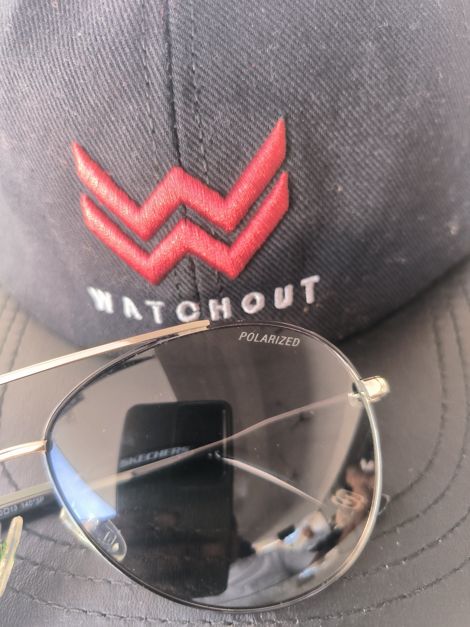 The image shows a black baseball cap with a red stylized "W" logo and the word "WATCHOUT" embroidered in white. A pair of gold-rimmed aviator sunglasses with "POLARIZED" printed on the lens is placed on top of the hat. The reflection in the sunglasses reveals a smartphone and a blurred figure. The scene appears to be shot outdoors, likely in Lhokseumawe, Indonesia, though there are no specific visual indicators to confirm this location within the image itself.