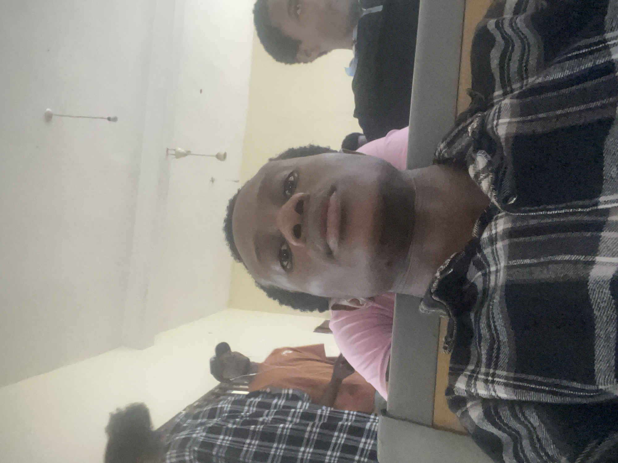 The image is a selfie taken indoors, likely in a classroom or lecture hall in Ihiagwa, Nigeria. The foreground is dominated by the face of a young Black man, who is looking directly at the camera. He is wearing a black and white checkered shirt. In the mid-ground, a person in a pink shirt is visible, and behind them, another person in an orange shirt wearing a black cap is looking to their left. The background shows a light-colored wall and ceiling, with two light fixtures hanging down. The overall lighting is bright and diffused.