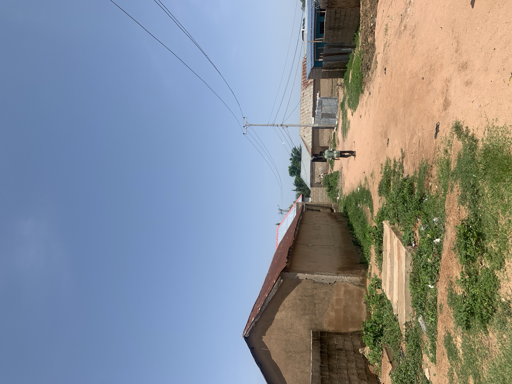 The image depicts a residential street in Jalingo, Nigeria. A person wearing a green and white patterned shirt and dark pants walks away from the camera down a dirt road. The road is flanked by simple, stucco-walled houses with corrugated metal roofs. Some of the houses have brick walls in front. There is vegetation growing along the sides of the road, and a concrete structure with several steps is visible on the left. Electrical poles and wires are present, indicating utility infrastructure. The sky is clear and blue.