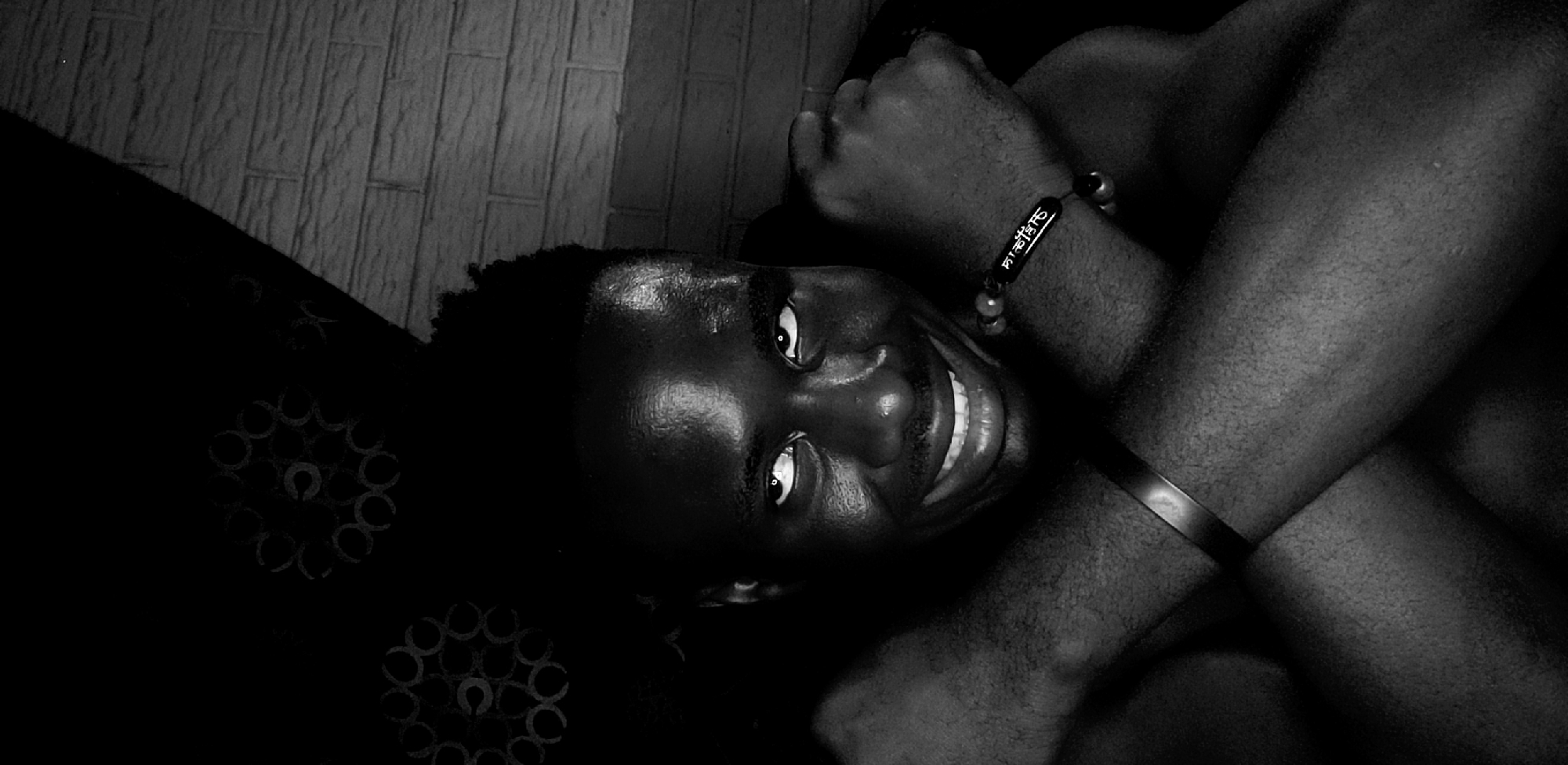 The image is a black and white photograph of a man with dark skin. He is lying down and looking up at the camera with a smile. His arms are crossed over his chest, and he is wearing a bracelet with an inscription on his left wrist. The location is Narayi, Nigeria. The background includes a patterned textile on the left and a tiled wall behind the man.