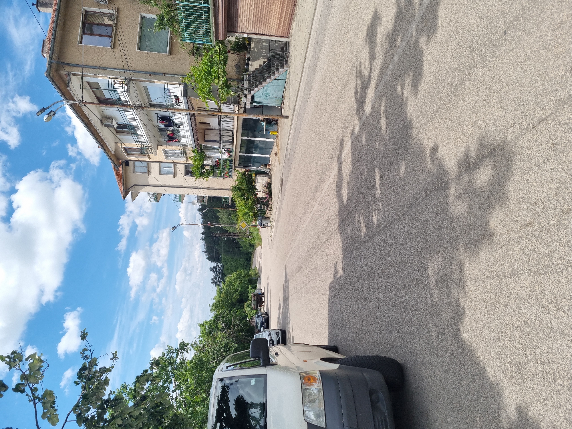A public road in Belogradchik, Bulgaria, curves towards the horizon, flanked by a multi-story building on the right and trees on the left. A white van is parked on the left side of the road. Several other vehicles, including a black car and a utility vehicle, are parked further down the road. The building on the right features balconies with laundry hanging out to dry and potted plants. The road is paved and shows shadows cast by trees. The sky is blue with scattered white clouds, and there are streetlights and utility poles along the roadside.