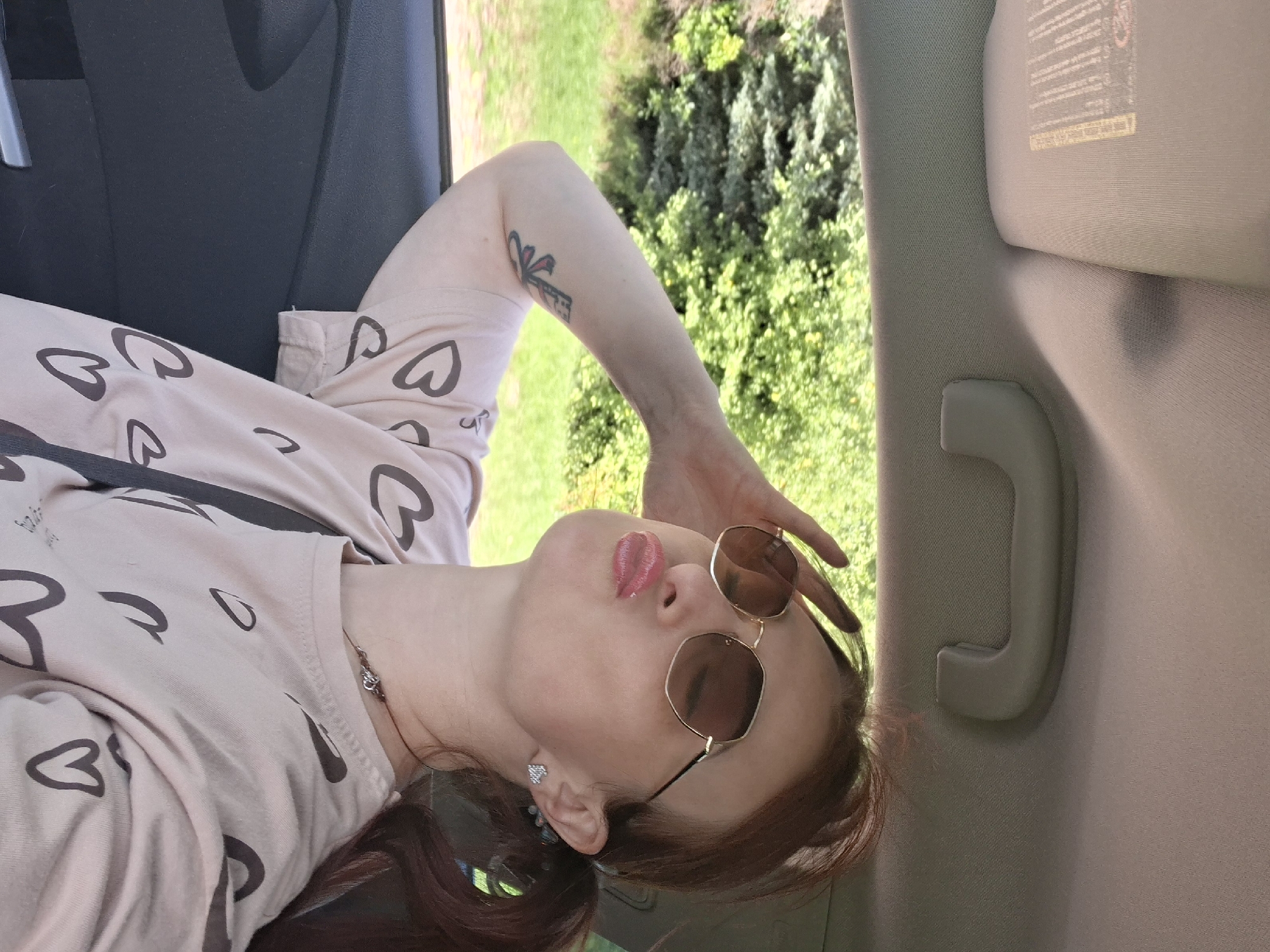 The image shows a young woman with auburn hair tied in a ponytail, wearing sunglasses and a light pink t-shirt with a heart pattern. She is inside a car, leaning her head back and holding her left hand to her forehead, with her lips pursed. The interior of the car is visible, including a grey door panel and part of the car's ceiling with a sun visor and a grab handle. Through the car window, lush green trees and vegetation are visible, suggesting an outdoor, possibly rural or suburban, setting. The presence of a car interior and the view of greenery from the window indicate the woman is a passenger in a vehicle moving through a natural landscape. No specific landmarks or features identifiable with the city of Myadzyel, Belarus, are present in the image.