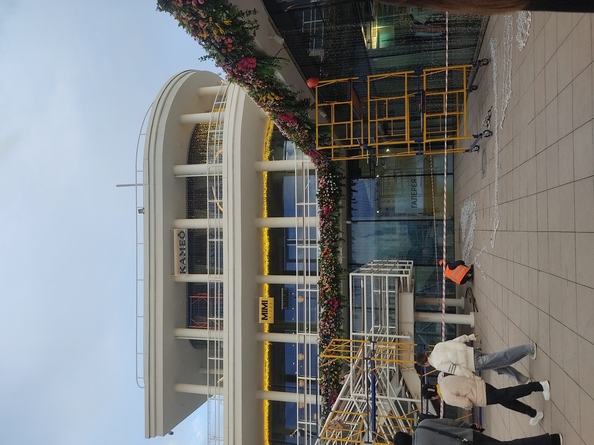 A ground-level shot presents a multi-story building with a curvilinear architectural design, adorned with a floral garland wrapping around the building’s facade. The structure has multiple levels with windows and lettering that reads "KAMBO" and "MIMI PIZZA" vertically. The building's reflective glass windows reflect light and possibly the surrounding environment.
Several yellow and white metal scaffolding structures are arranged in front of the building. People are visible in the foreground, some appearing to walk past the scene and others working near the scaffolding. One individual is wearing a bright orange vest.
White decorative string lights or garlands are draped or hanging near the edge of the building. The sky appears overcast and light.
Based on the Cyrillic lettering "ГАЛЕРЕЯ" (Gallery) visible on a building sign, the location may be in Russia, potentially Moscow.