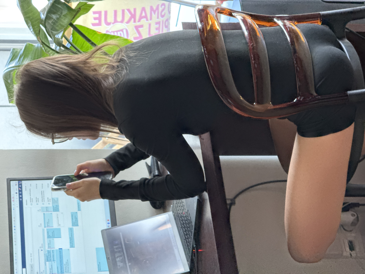 The image depicts an indoor scene in what appears to be an office, co-working space, or cafe in Warsaw, Poland, during daytime. The central subject is a person, likely a woman, with long brown hair, wearing glasses and a black long-sleeved top or bodysuit. Her back is partially towards the viewer, and she is leaning over a desk or table, focused intently on a smartphone held in both hands.
On the desk in front of her is a computer monitor displaying what appears to be a scheduling or task management interface with various blue and white boxes. An open laptop is positioned to the right of the monitor. The woman's lower body, specifically her buttocks and upper thigh, is visible between the distinctive amber-colored, curved bars of a wooden chair's backrest and its seat. Her right leg is partially visible below the chair, bent at the knee.
In the upper left background, a large green leafy plant is visible against a window or glass partition. This glass reflects mirrored text, which reads "SNAWKAME" (likely "EMAKWANS" or similar when unreversed) and the numbers "1319" (possibly "9131" or part of a phone number/address when unreversed), suggesting signage from outside or within the establishment. The woman seems engaged in a task, possibly interacting with her phone while near her workstation.