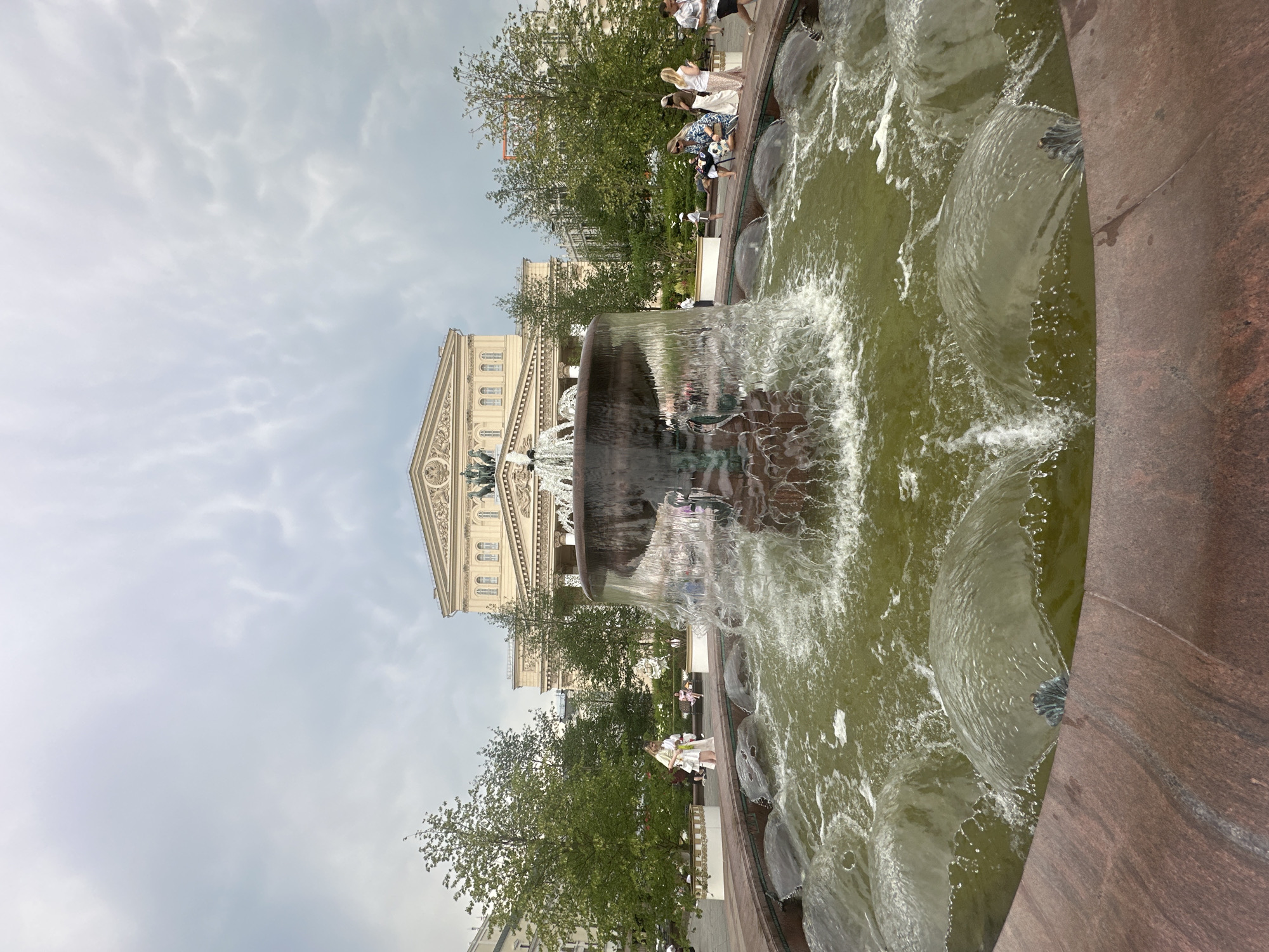 The image shows a fountain in front of a grand building, believed to be the Bolshoi Theatre in Moscow, Russia, under a cloudy sky. The fountain is active, with water splashing and creating a mist. Several people are visible in the background, walking and sitting around the fountain. The architecture of the building is classical, with columns and intricate detailing. Trees and greenery surround the area, suggesting a public square or park. The overall atmosphere appears to be leisurely, with people enjoying the outdoor space.