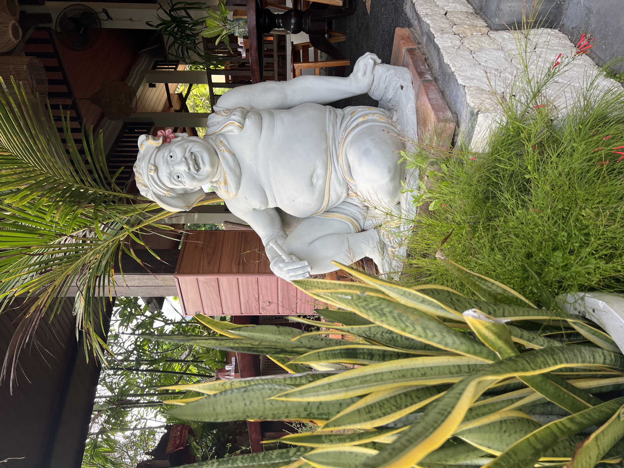 The image displays an outdoor scene centered around a white stone statue of a jovial figure. The statue depicts a portly man with a wide, open-mouthed smile, large expressive eyes, and a small pink flower tucked behind his left ear. Subtle golden accents adorn his clothing, suggesting decorative patterns. He is seated with bent legs and hands resting on his thighs, conveying an emotion of cheerfulness.
The statue is positioned on a raised base made of light-colored, rough-textured stones, with some reddish bricks visible nearby. Lush tropical greenery surrounds the figure. In the foreground on the left, a large snake plant (*Sansevieria trifasciata*) with distinctive green and yellow striped leaves dominates. To the right of the statue, a bushy plant featuring slender green stems and small red tubular flowers, possibly a firecracker plant, adds a vibrant touch.
In the background, a building with wooden architecture is visible, likely a restaurant or cafe. It features wooden walls, pillars, and roof elements, along with rattan decorations like hanging light fixtures and baskets. Dark wooden tables and chairs are arranged within the covered area. Overhead, large palm fronds extend into the frame from the upper left. The natural lighting suggests daytime, possibly late morning or early afternoon, with soft shadows. The overall setting is lush and tropical, consistent with the location context of Kuta Selatan, Indonesia, and evokes a relaxed, culturally infused atmosphere. There is no visible text in the image.