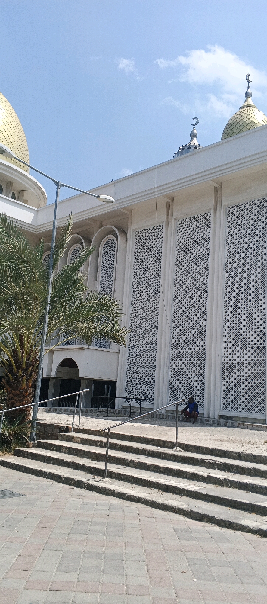 The image shows a wide shot of the exterior of a mosque with a golden dome, possibly located in Kandangsemangkon, Indonesia. The mosque features white walls with intricate geometric patterns in its windows. In the foreground, there are wide stone steps leading up to the entrance, with metal railings on either side. To the left, a palm tree with green fronds is visible. A person is sitting on the steps near the base of the mosque, dressed in blue and red clothing. The sky above is clear blue with a few wispy clouds. The overall scene suggests a bright, sunny day.
