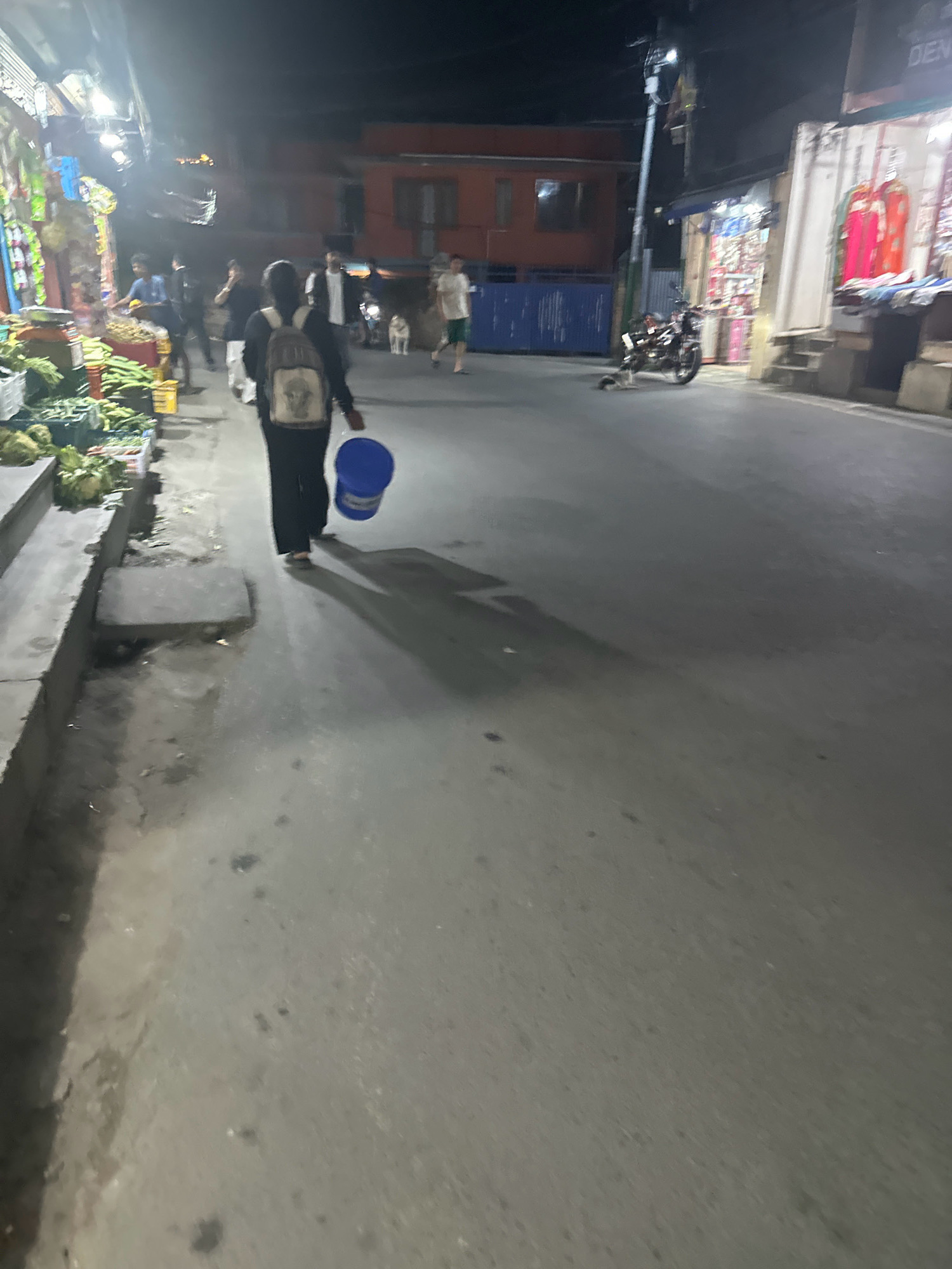The image depicts a street scene at night in Lalitpur, Nepal. A person, seen from behind, walks away from the camera carrying a blue bucket. The person is wearing a backpack. To the left, there are stalls with produce, likely a market. In the background, there are buildings, a motorcycle, and other individuals. The street is paved, and lighting appears to be from street lamps and shop displays. The overall atmosphere is that of a bustling urban environment after dark.