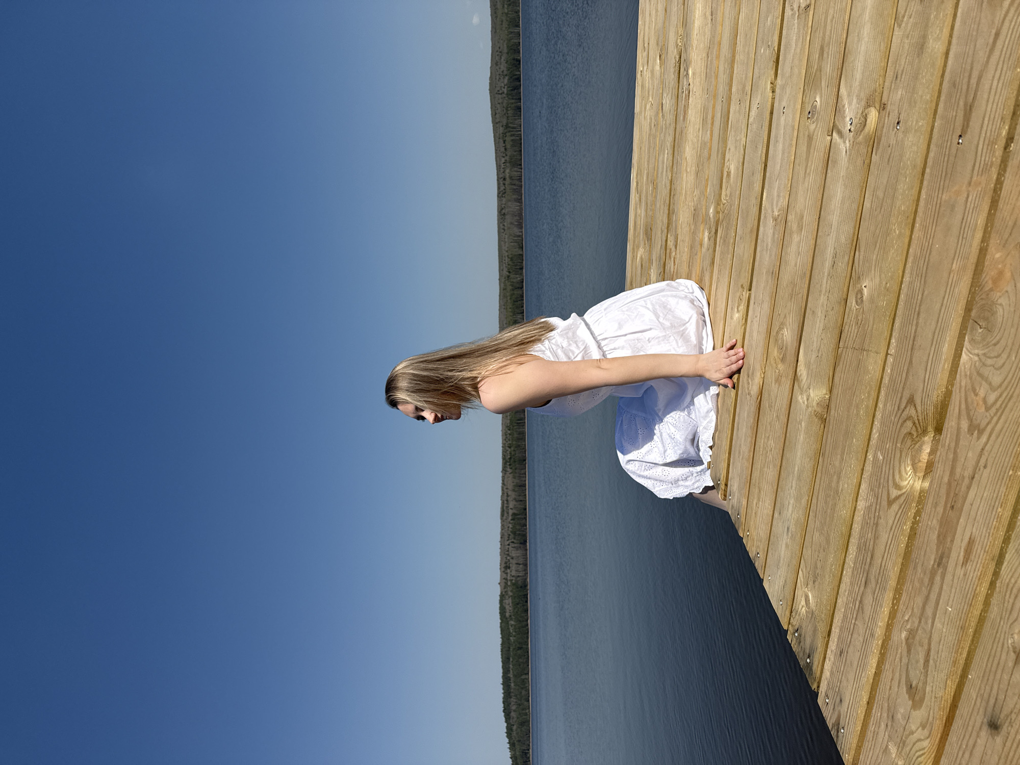 A young woman with long blonde hair, wearing a white dress, kneels on the edge of a wooden dock and peers into the water. The dock extends into a body of water, possibly a lake or large river, with a distant shoreline visible. The sky is clear and blue. The scene appears to be set in a natural environment, possibly near the city of Chaikovsky, Russia, given the prompt.