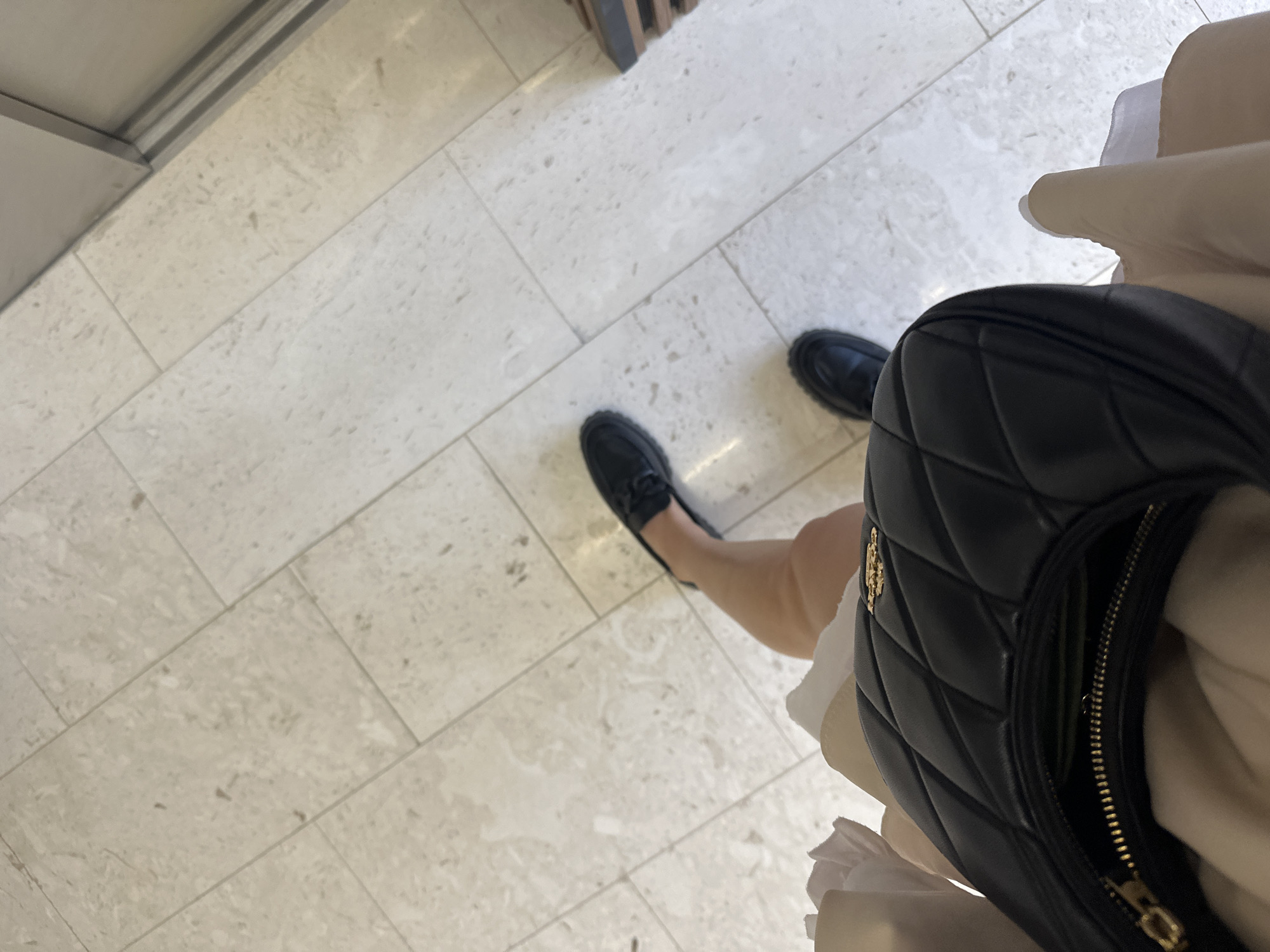 From a high-angle perspective, the image displays a portion of a person standing on a light beige, patterned tile floor. The individual is wearing black loafers and a light-colored dress. A black quilted bag is positioned in the foreground, partially obscuring the view. The location cannot be determined solely from the provided image. The scene appears to be indoors.
