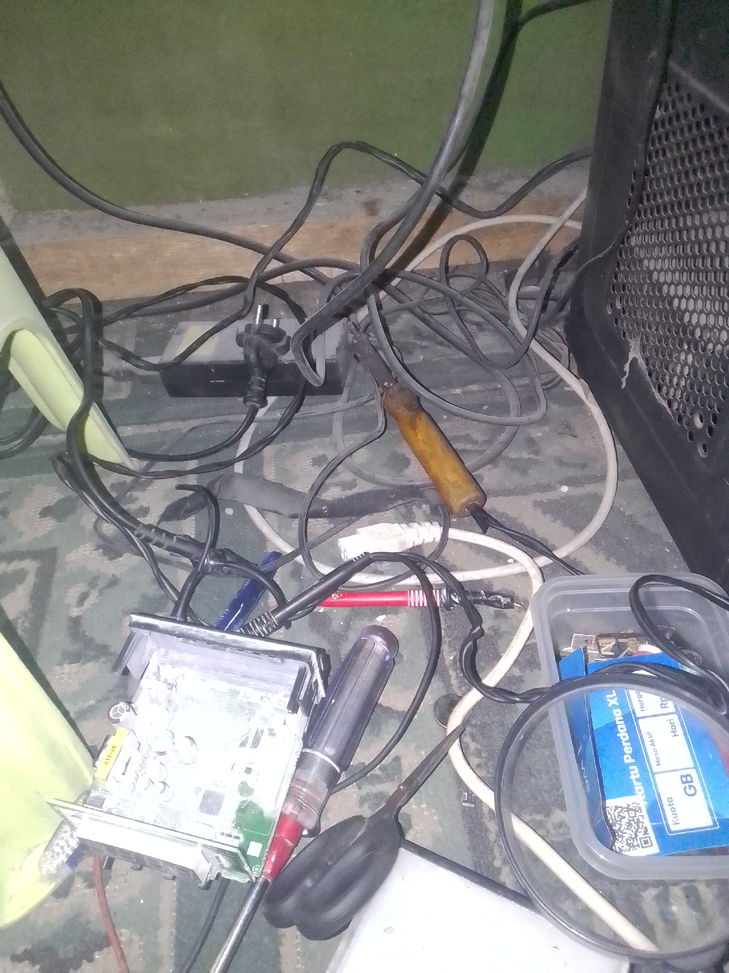 A cluttered workspace is shown in Gunungmasigit, Indonesia. A soldering iron with a wooden handle rests on a tangle of black and white wires. A circuit board, possibly from an electronic device, is partially visible in the foreground, connected to wires. To the right, a clear plastic container holds various small electronic components and a blue card with text in Indonesian. A magnifying glass is positioned over the container. A black speaker cabinet is visible on the right side of the image. The overall impression is one of an active but disorganized electronic repair or hobbyist area.
