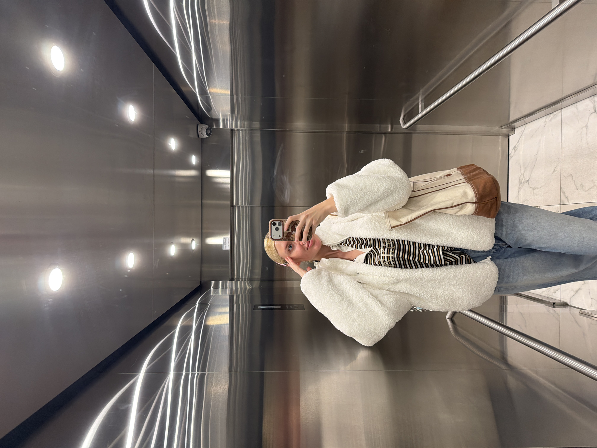 A woman with blonde hair is taking a selfie in an elevator. She is wearing a white, fuzzy jacket, a striped shirt, and blue jeans. She is holding a smartphone in her right hand. Behind her, the elevator walls are made of shiny metal with evenly spaced inset lighting. A security camera is also visible in the upper portion of the elevator. A handrail runs along one side. The elevator floor appears to be marble. The location in the city of Belgrade, Serbia, is not discernible from the image.