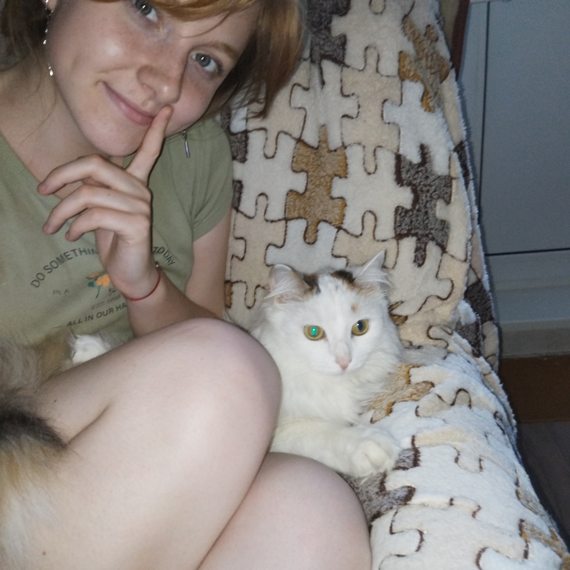 A young woman with reddish-blonde hair is sitting on a patterned couch with a white and tan cat. The woman is wearing a light green t-shirt with text and an illustration. She is holding a finger to her lips. The cat is mostly white with some tan markings on its head and back. The couch has a pattern of interlocking puzzle pieces in white, tan, and brown. A white door frame is partially visible in the background. The specific location within Yekaterinburg, Russia cannot be determined from the visual data alone.
