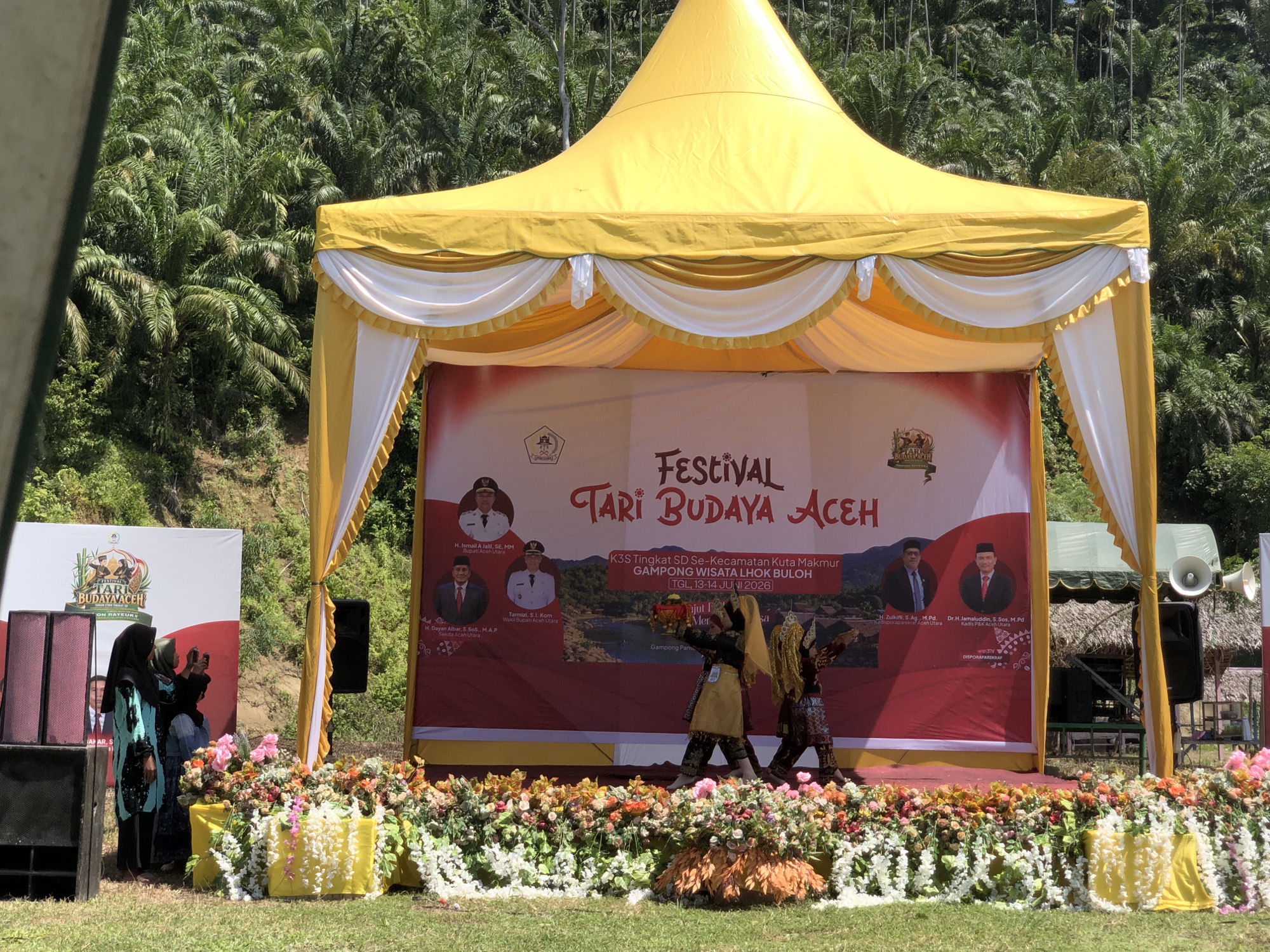 The image depicts an outdoor stage set up for a cultural festival, likely in the city of Langkuta, Indonesia, given the banner text. A large yellow tent with white draping covers the stage. In the background, a lush green hillside covered in palm trees suggests a tropical environment. The stage itself is adorned with a colorful floral arrangement in the foreground.
A large banner on the stage reads "FESTIVAL TARI BUDAYA ACEH," indicating a festival of Acehnese dance culture. The banner also contains text about the event's organization: "K3S Tingkat SD Se-Kecamatan Kuta Makmur Gampong Wisata Lhok Buloh," with dates "TGL 13-14 JUNI 2026." Several portraits of men in formal attire are visible on the banner, along with their names and titles, including "Bupati Aceh Utara" and "Wakil Bupati Aceh Utara."
On the stage, two individuals in traditional attire are performing a dance. One is holding a prop. Two women in the foreground, dressed in headscarves and modest clothing, are observing the performance, with one appearing to take a photo with her phone. Two large black speakers are positioned on either side of the stage. The overall scene is brightly lit by natural sunlight.
