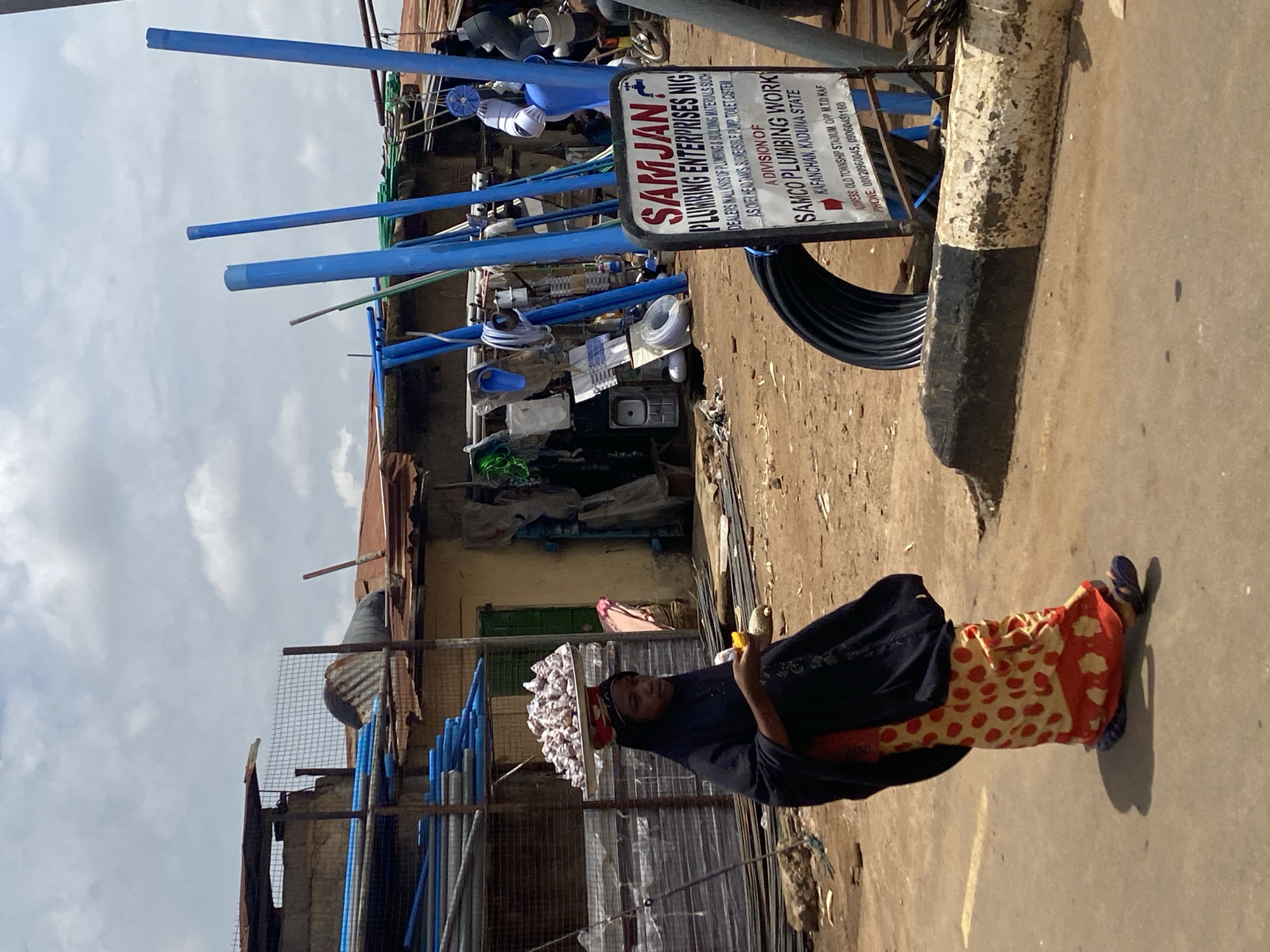 The image, rotated vertically, depicts an outdoor street scene in Kafanchan, Kaduna State, Nigeria, under a bright, partly cloudy sky.
In the foreground, a woman is seen walking towards the left. She is carrying a flat tray piled with numerous small, light-colored items, possibly garlic or similar produce, on her head. She wears a dark, long-sleeved top, a vibrant red and yellow polka-dotted skirt, and blue flip-flops. She holds a yellow object in her right hand, and her expression is neutral.
Behind the woman, to the right, is what appears to be a plumbing supply shop. Various plumbing materials are displayed, including numerous blue and grey pipes, large black corrugated pipes, sinks, fittings, and other components. A prominent white sign with red lettering stands upright in front of the shop.
The visible text on the sign reads: "SAMJAN PLUMBING ENTERPRISES NIG," "DEALERS WALL INIUS OF PLUMBING & BUILDING MATERIALS SUCH AS OVER HEAD TANKS SUBMERSIBLE PUMP TOLET COSTEM," "A DIVISION OF SAMCO PLUMBING WORK," "KAFANCHAN, KADUNA STATE," "ADRESS, OLD TOWNSHIP STADIUM OPP MTD KAF," and lists phone numbers: "08020960045, 09060451160."
The background features simple, weathered buildings with corrugated metal roofs and metal grating. A black and white painted concrete bollard is partially visible on the far right. The ground is an unpaved or lightly paved surface. The scene portrays a typical daytime street environment with local commerce.