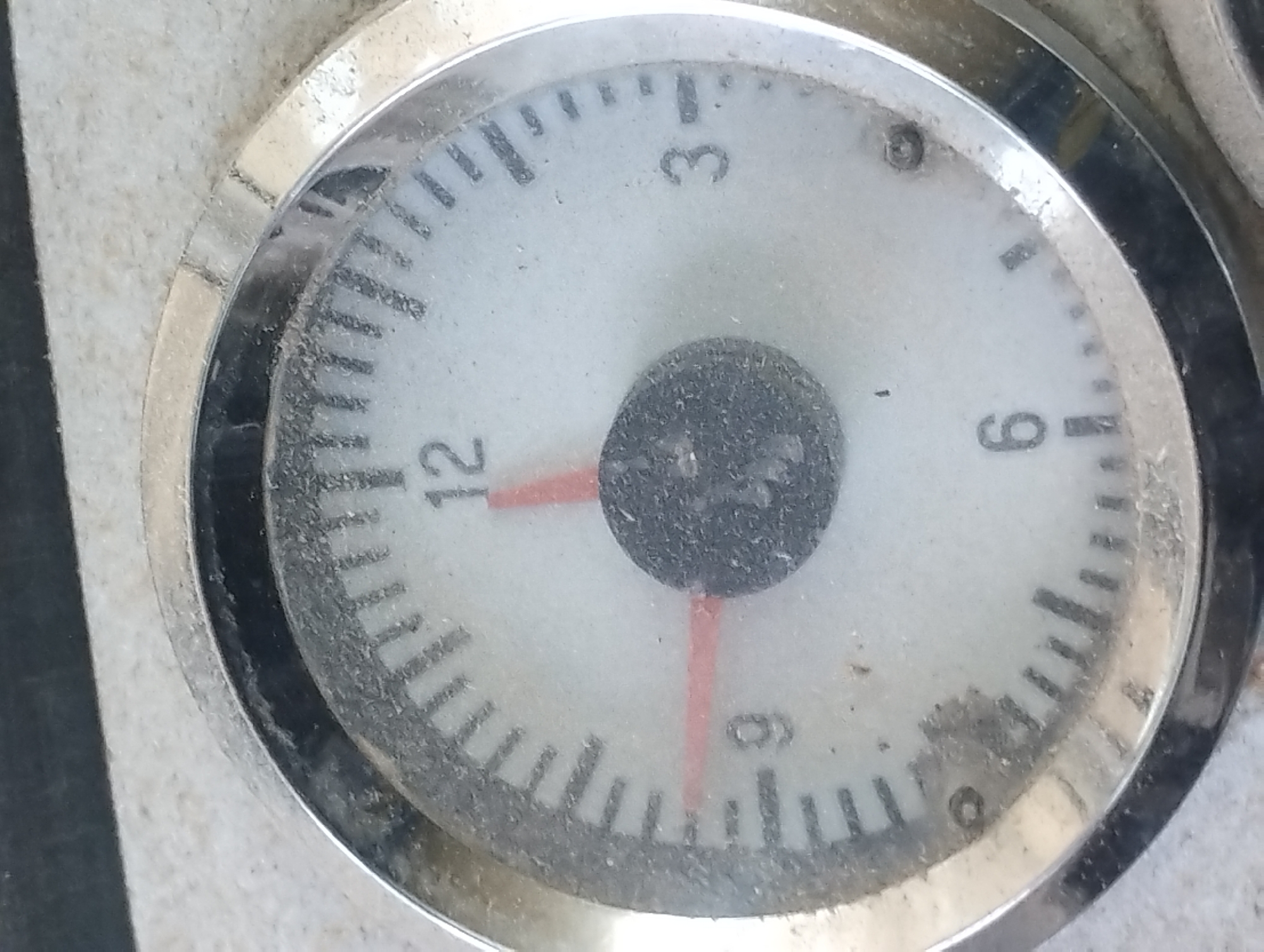 The image shows a close-up of a dusty analog clock face. The clock has a white face with black hour markers and numbers. The numbers visible are 12, 3, 6, and 9. There are two hands on the clock: a shorter, thicker red hand pointing roughly towards the 9, and a longer, thinner red hand pointing roughly towards the 12. The clock is housed in a silver bezel. The background is out of focus and appears to be a textured, neutral-colored surface. There is no discernible location in the city of Sredets, Bulgaria visible in this image.