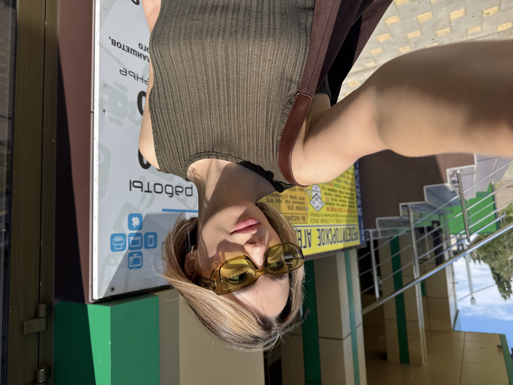 A young woman with blonde hair and sunglasses is taking a selfie outdoors in front of a building. She is wearing a ribbed, olive-green tank top and a brown strap is visible over her shoulder from a bag. Behind her, there is a building with green and beige accents, a staircase with a silver railing, and a sign with various icons, including a computer, a phone, and an apple logo. The ground is paved with light-colored square tiles. The overall lighting suggests it is a sunny day.