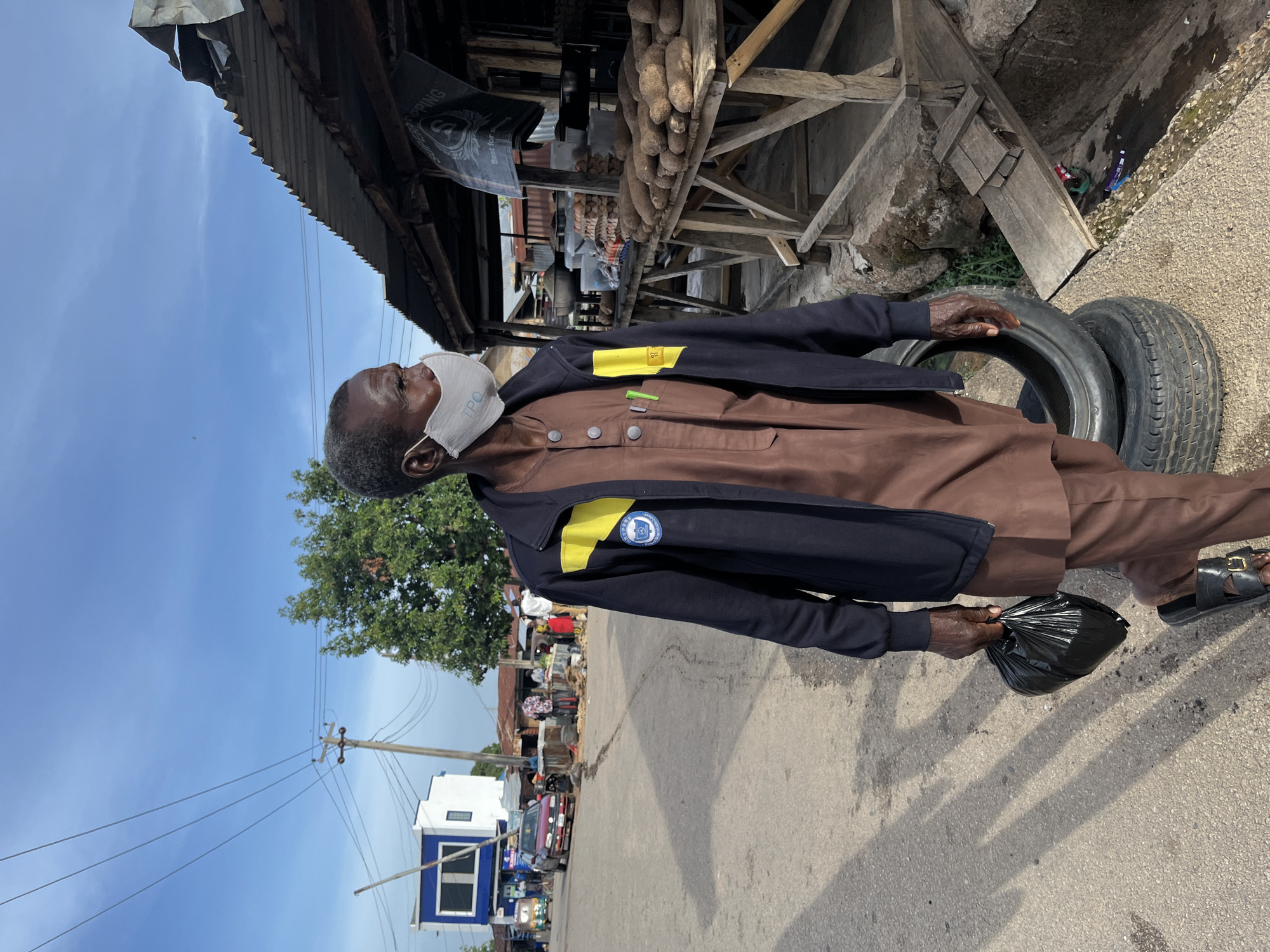 The image, taken in Jos, Nigeria, depicts an elderly man standing on a paved road under a clear, bright blue sky, indicative of midday or early afternoon. He is dressed in a brown traditional tunic and trousers, topped with a dark blue jacket featuring a yellow stripe and a circular logo on the left arm. A white face mask, partially revealing "TPO" text, covers his mouth and nose. The man holds a black plastic bag in his left hand and looks upwards and slightly to his left.
To the man's immediate right, two old car tires rest on the ground next to a rustic, makeshift market stall. This stall features a corrugated metal roof supported by wooden beams, and beneath it, numerous yams or similar root vegetables are displayed on a wooden stand. A dark bag hanging in the background shows partial text: "STOPPING" and "Best for".
The background on the left side of the image reveals a vibrant street scene characteristic of a market. Various stalls, some people, and hints of vehicles are visible. Overhead, power lines crisscross the sky, leading to a utility pole. Further down the street, a distinctive blue and white building stands out. The overall atmosphere suggests a busy, outdoor commercial area.
