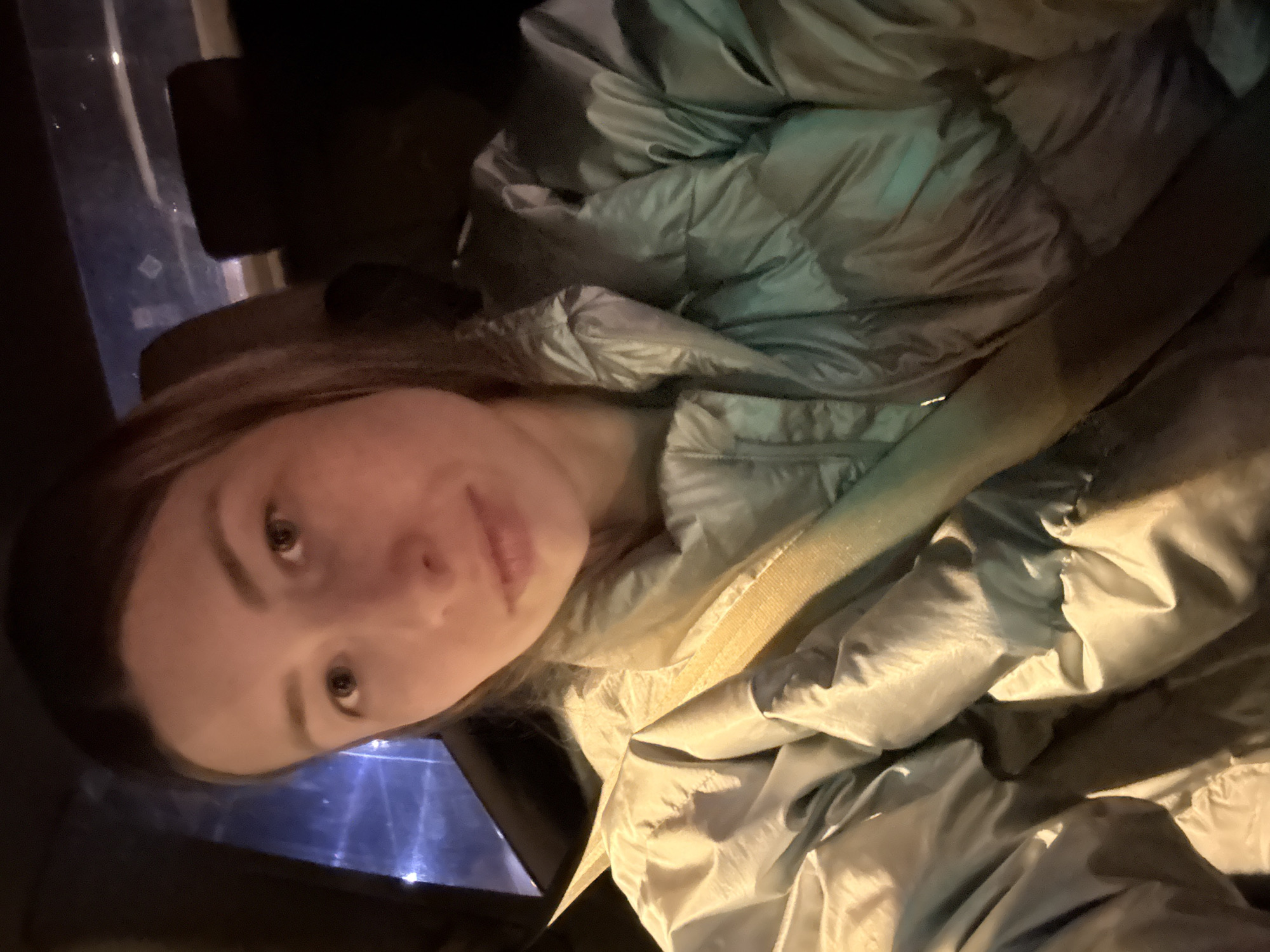 A female with medium-length brown hair and light skin looks directly at the camera. She is wearing a shiny, light blue puffer jacket. The setting appears to be inside a vehicle at night, with a faint blue light emanating from the window behind and to the left of her. The location is not discernable as Moscow or any other specific city.
