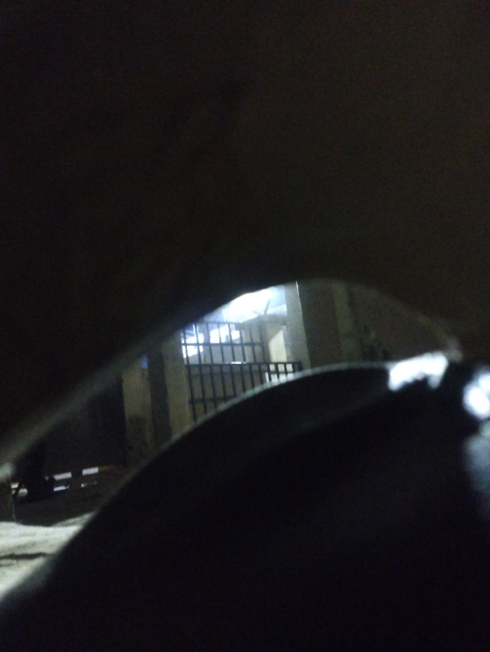 The scene, located in Kubare, Nigeria, is captured at night, characterized by low light conditions with a bright artificial light source in the background. The foreground features a dark, curved object, likely an internal component of a vehicle, which partially frames the central view. Through this opening, a metal gate with vertical bars is visible. Behind the gate, structural pillars of a building are present, beyond which the bright light source illuminates an area. To the left of the gate, two distinct, parallel white horizontal markings are discernible on a dark, level surface. No individuals are depicted, and no specific actions or interactions are occurring. The visible environment includes paved ground.