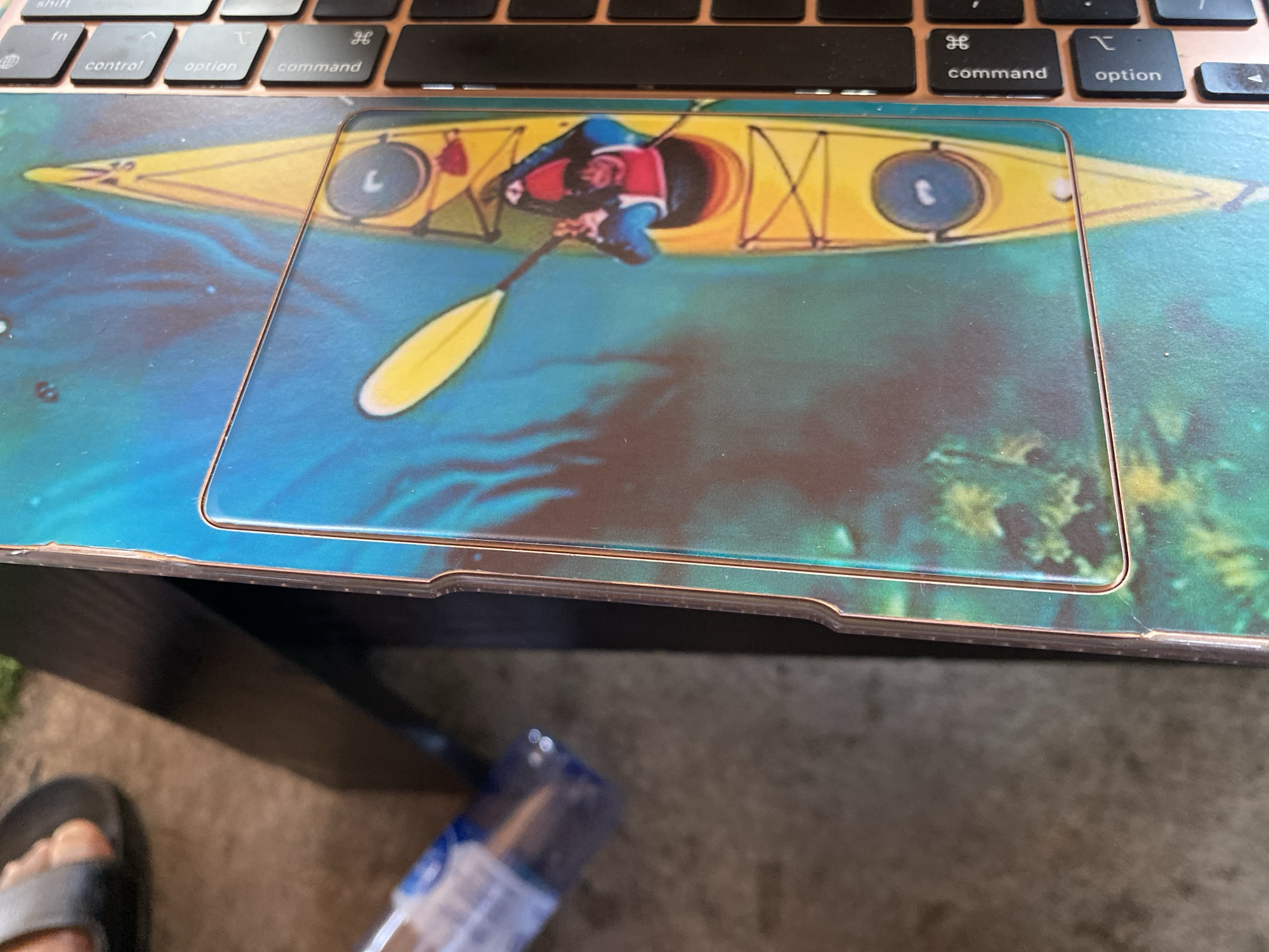 The image shows a close-up of a laptop with a custom skin featuring a yellow kayak with a person paddling. The kayak is positioned on a blue and green watery background. Part of a hand and foot wearing a sandal are visible in the upper left corner. The laptop keyboard is visible on the right side of the image, with keys labeled "control," "option," and "command." The scene appears to be indoors, possibly in a home or office setting, given the presence of a laptop and furniture. The image does not contain information to determine its location in Howayi, Nigeria.