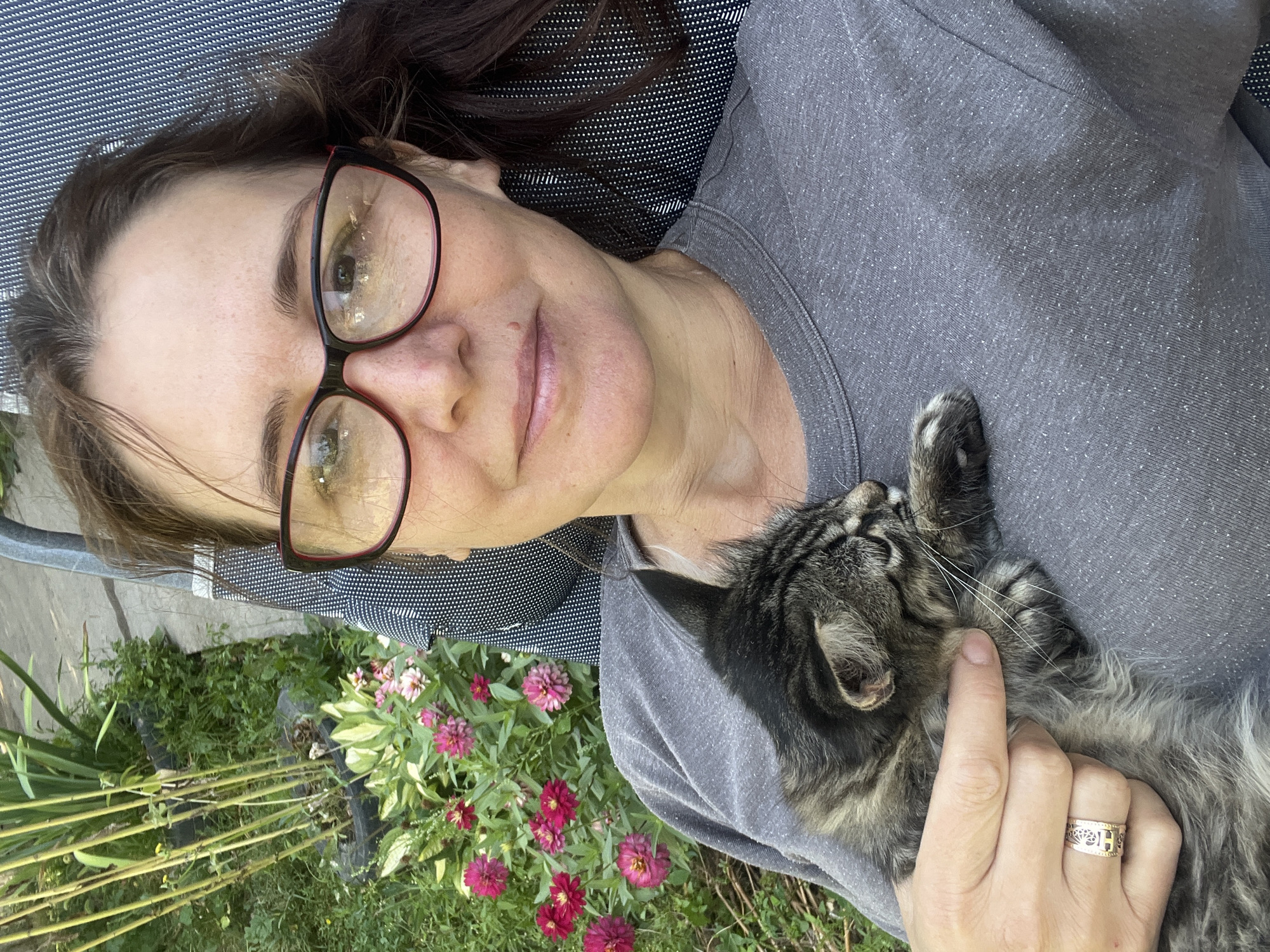 A woman with medium-length brown hair, wearing glasses with red frames and a grey t-shirt, is holding a tabby cat in her arms while lying down outdoors. She is wearing a ring on her left hand with a Celtic knot design. Behind her, there is a garden with various plants and flowers, including tall green stalks and pink and red zinnias. The setting appears to be a backyard or park in Қарғалы, Kazakhstan. The woman is looking towards the camera, and the cat is nestled comfortably in her arms. The overall impression is one of a relaxed, domestic scene.