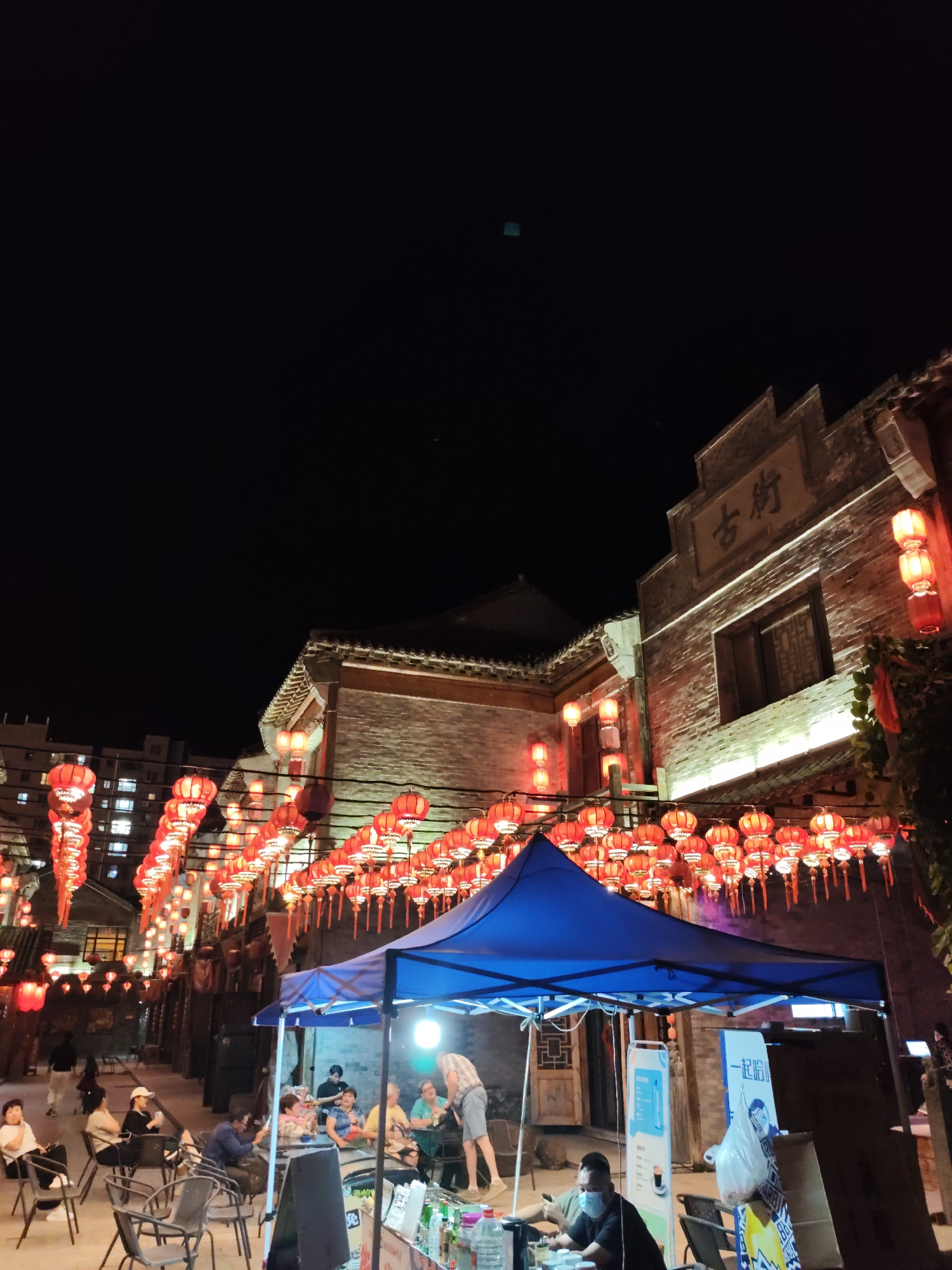 This image depicts an outdoor night scene in Lindian, China, likely a pedestrian street or market area decorated with numerous red lanterns strung across the pathway. Buildings with traditional Chinese architectural elements, constructed from brick, flank the street. One building on the right has a sign with Chinese characters that appear to read "古街" (Gu Jie), meaning "Ancient Street."
In the foreground, a blue pop-up tent covers a small food or drink stall, suggesting commercial activity. Several people are seated at tables and chairs under or near the tent, some engaged in conversation or looking at their phones. A person wearing a mask is seen at the stall.
The street is illuminated by the red lanterns, the lights from the buildings, and spotlights illuminating the lower portions of the brick walls. In the background, a modern apartment building is visible, indicating the coexistence of old and new architecture. The sky is dark, typical of nighttime. The overall atmosphere suggests a lively evening gathering or market.