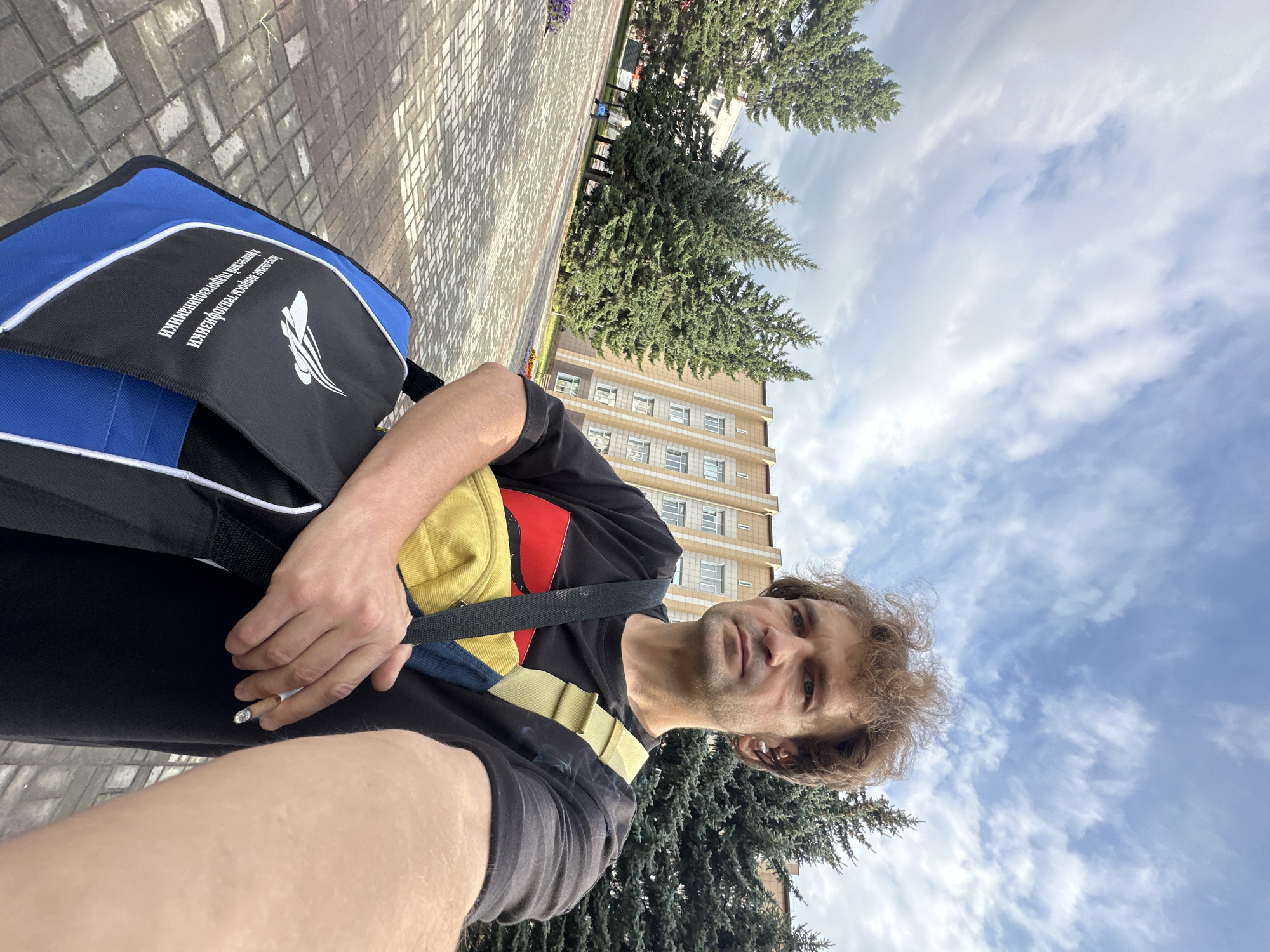 A man stands outside, taking a selfie in front of a building in Biysk, Russia. He has curly brown hair, a serious expression, and is wearing a black t-shirt with red and yellow accents. He has a yellow and blue bag slung across his body and is holding a lit cigarette in his right hand. The background shows a paved courtyard, several evergreen trees, and a tan building with a repeating window pattern. The sky is cloudy with patches of blue.
