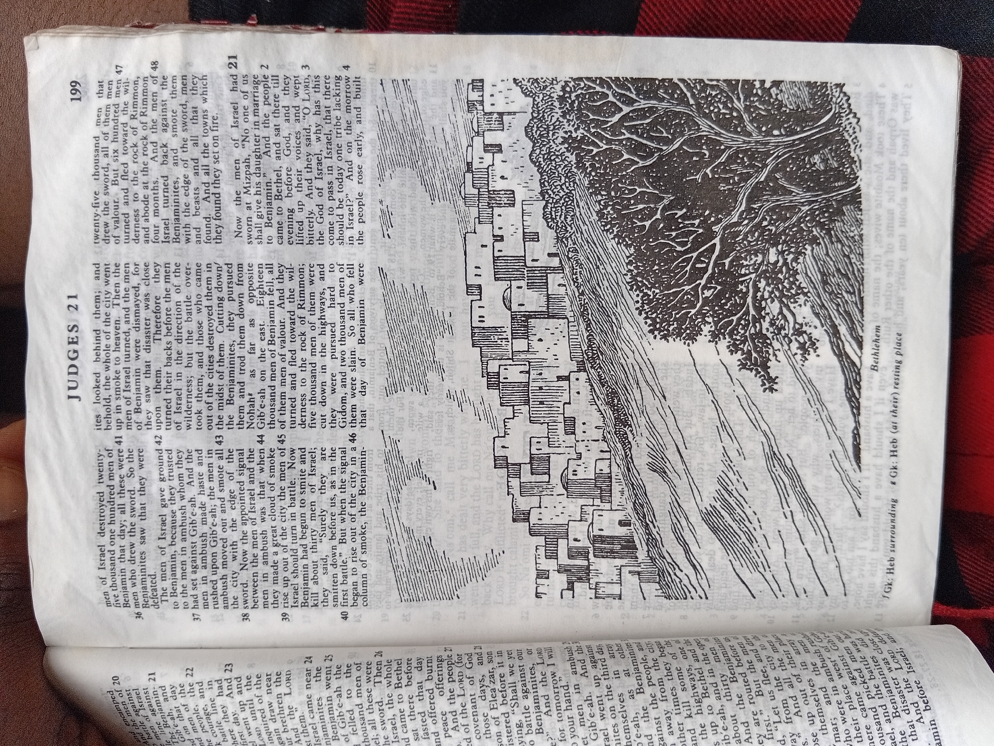The image displays an open book, revealing a page of text and an illustration. The text is in English and appears to be from the Book of Judges, specifically chapter 21, with verse numbers indicated. The illustration is a woodcut-style drawing depicting a city with many small, uniform buildings and a prominent tree to the right. The page also includes what appears to be a caption in a smaller font underneath the illustration. There are no people or identifiable subjects depicted in the illustration. The book is placed on a surface with a red and black checkered pattern. No information about the city of Nkpor, Nigeria, is present in the image.
