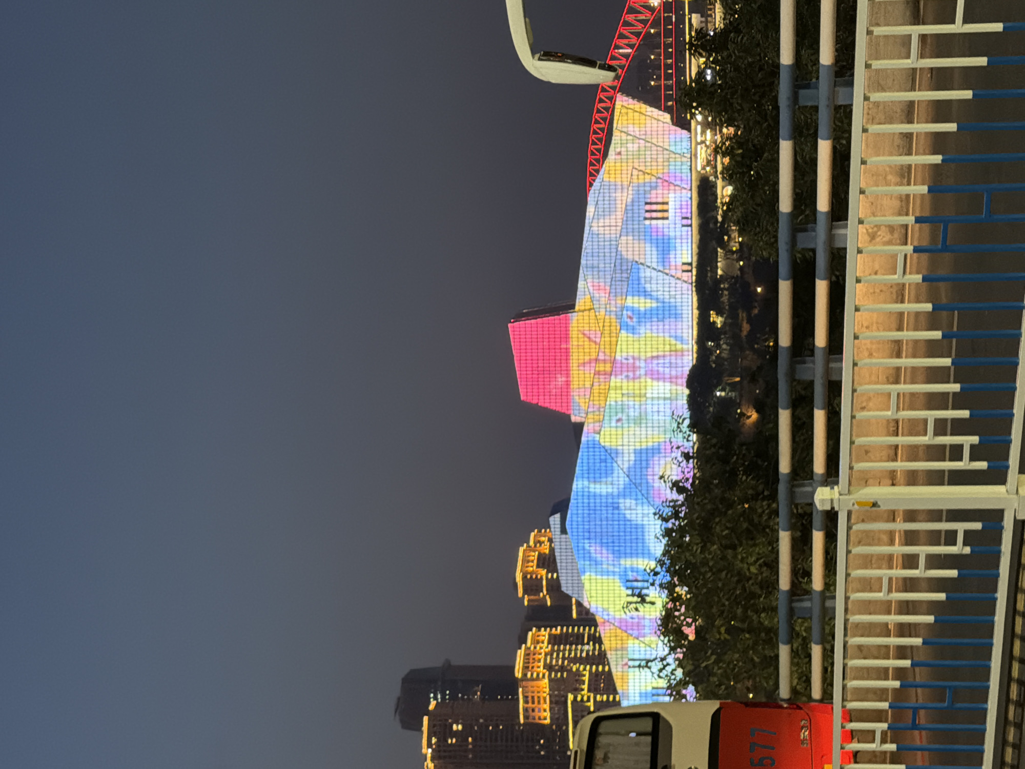 In Yuzhong District, China, at twilight, a striking modern building with a multi-colored, illuminated facade is visible. The building's exterior displays a vibrant, abstract pattern of blues, yellows, and pinks, with some areas appearing to depict stylized figures. To the left of this building, several high-rise structures are illuminated with orange lights. A red bus with the number "577" is parked in the foreground, partially obscuring a dense green foliage. A white guardrail with blue and white horizontal bars runs across the bottom of the image, separating the viewer from the street or walkway. The sky is a dark, dusky blue, indicating the transition from day to night. A section of a red, illuminated bridge structure is visible to the right of the main building, and a white overhead structure, possibly part of a cable car or elevated walkway, is also present on the far right.