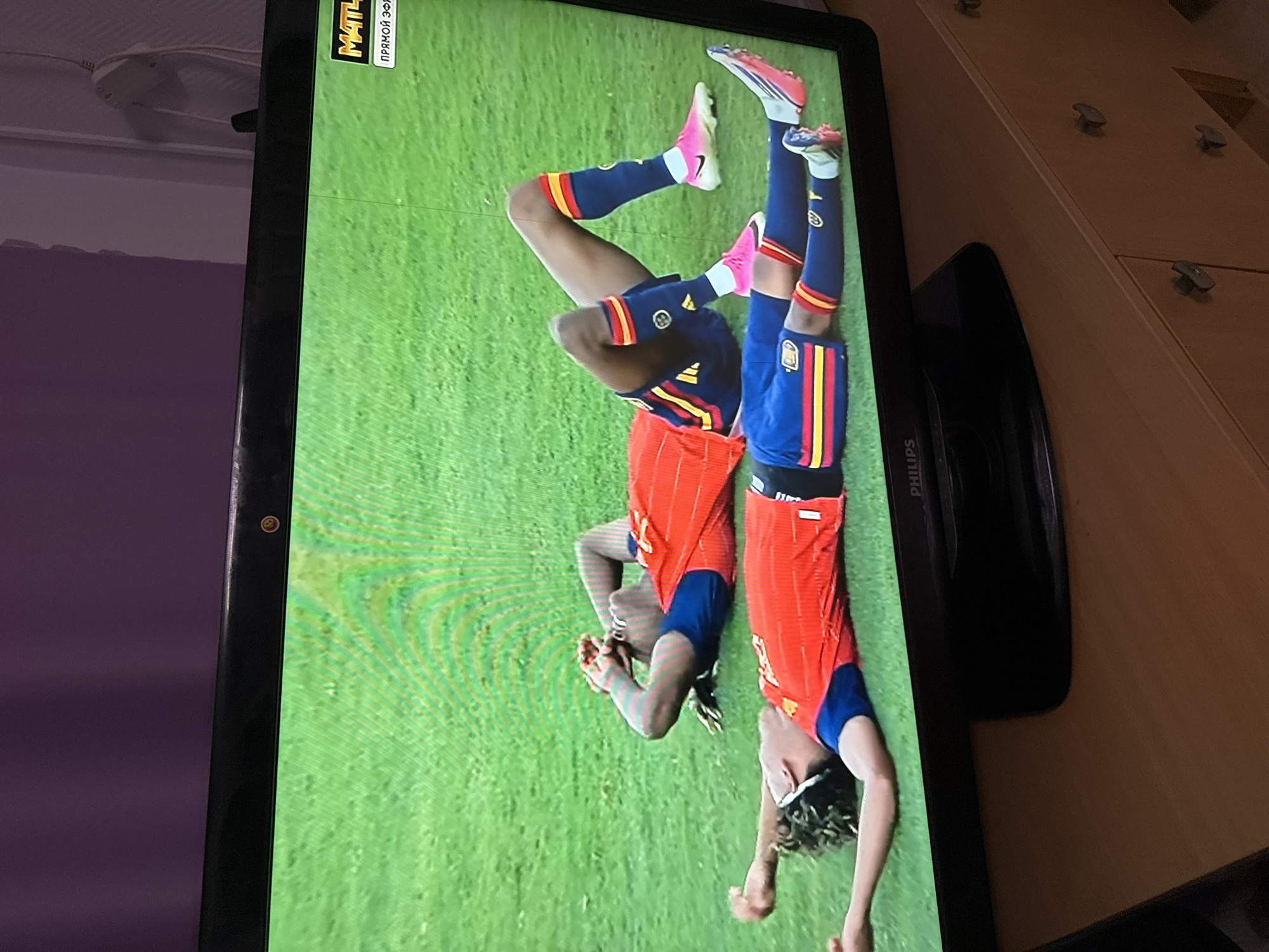Two soccer players in red and blue uniforms lie on a green grass field, seemingly exhausted. One player, in the foreground, is lying on his back with his arms outstretched. The second player, in the background, is lying on his side with his hands over his face. Both players have red and yellow striped socks and blue shorts with similar stripes. The image is a photograph taken of a television screen, indicated by the reflection on the screen and the visible edge of the television frame. A logo for "МАТЧ" (Match) and "ПРЯМОЙ ЭФИР" (Live Broadcast) is visible in the top right corner of the screen. The television is placed on a wooden surface, possibly a cabinet, with drawers and handles. The wall behind the television is a muted purple color. The context of Ufa, Russia, is not discernible from the visual information within the image itself.