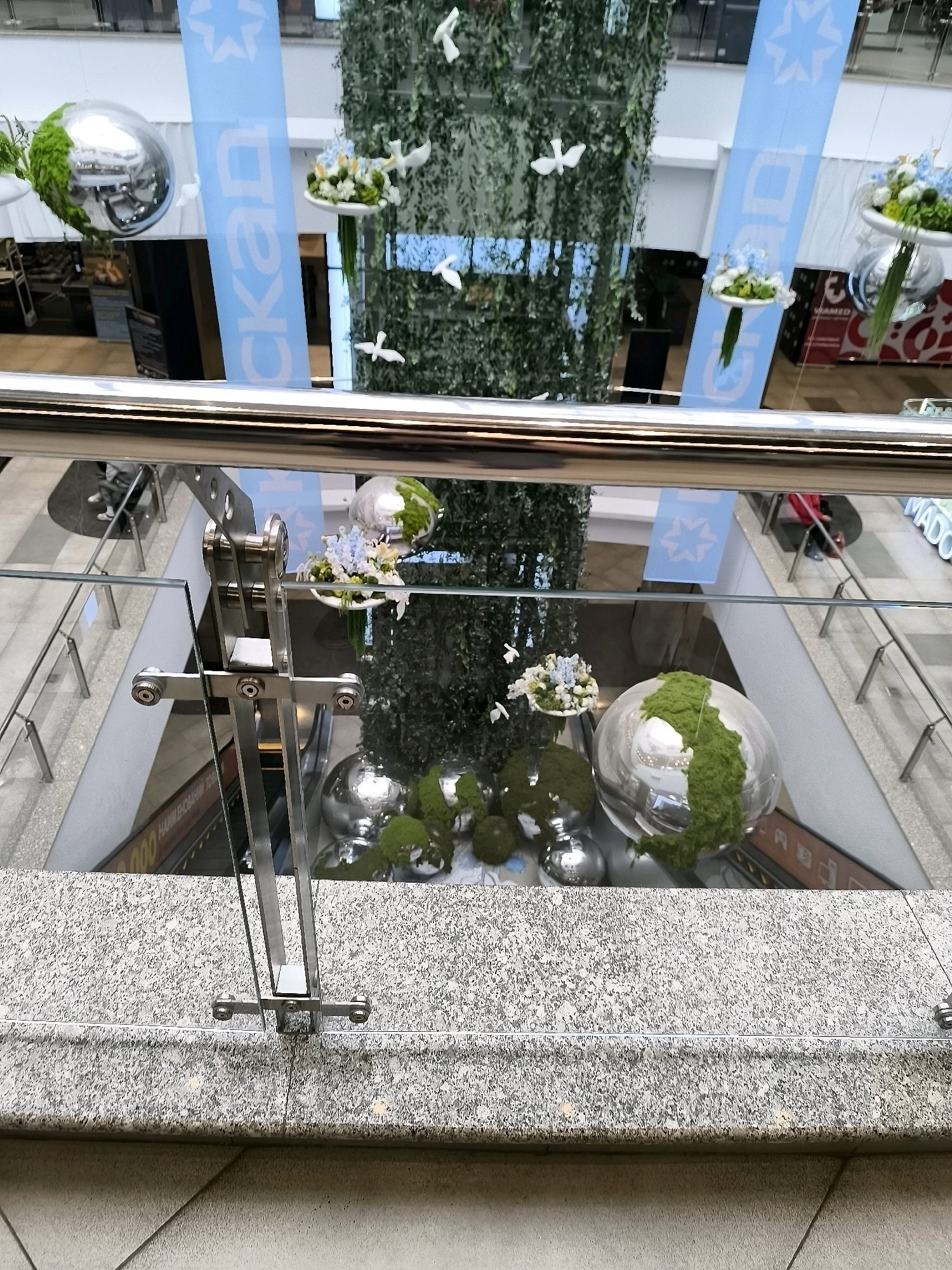The image shows an interior atrium of a multi-story building, likely a shopping mall or office complex. The focus is on a central column adorned with lush green vines and hanging decorations. Suspended from the column are several shiny, spherical objects, some partially covered in green moss-like material, resembling planets or decorative orbs. White dove-shaped ornaments are also scattered around the column, adding to a decorative theme.
In the background, a large blue banner with white lettering and a stylized star logo is visible. Glass railings and floors suggest multiple levels within the atrium. The lighting appears to be artificial, typical of an indoor commercial space.
While the image depicts an interior scene with decorative elements, there is no indication or identifiable context to suggest it is located in the city administration of Qarağandı, Kazakhstan. The environment appears to be a modern commercial or public indoor space.