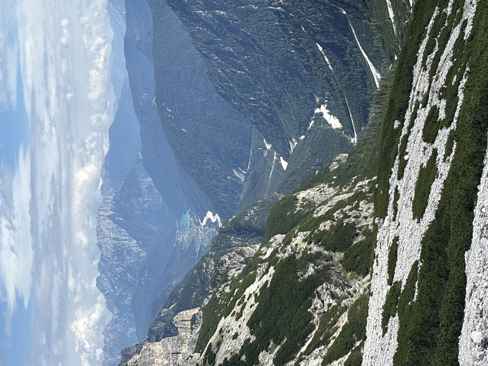 The image depicts a mountainous landscape in the vicinity of Auronzo di Cadore, Italy. The scene is dominated by rugged, rocky peaks with sparse vegetation, contrasting with the dense green forests covering the lower slopes. A winding river or road is visible in the valley below, with a small settlement nestled beside it. The sky is partly cloudy, with patches of blue sky visible between the clouds. The perspective is from a high vantage point, looking down into the valley and across the mountain range. The overall impression is one of vast, natural beauty and remote wilderness.