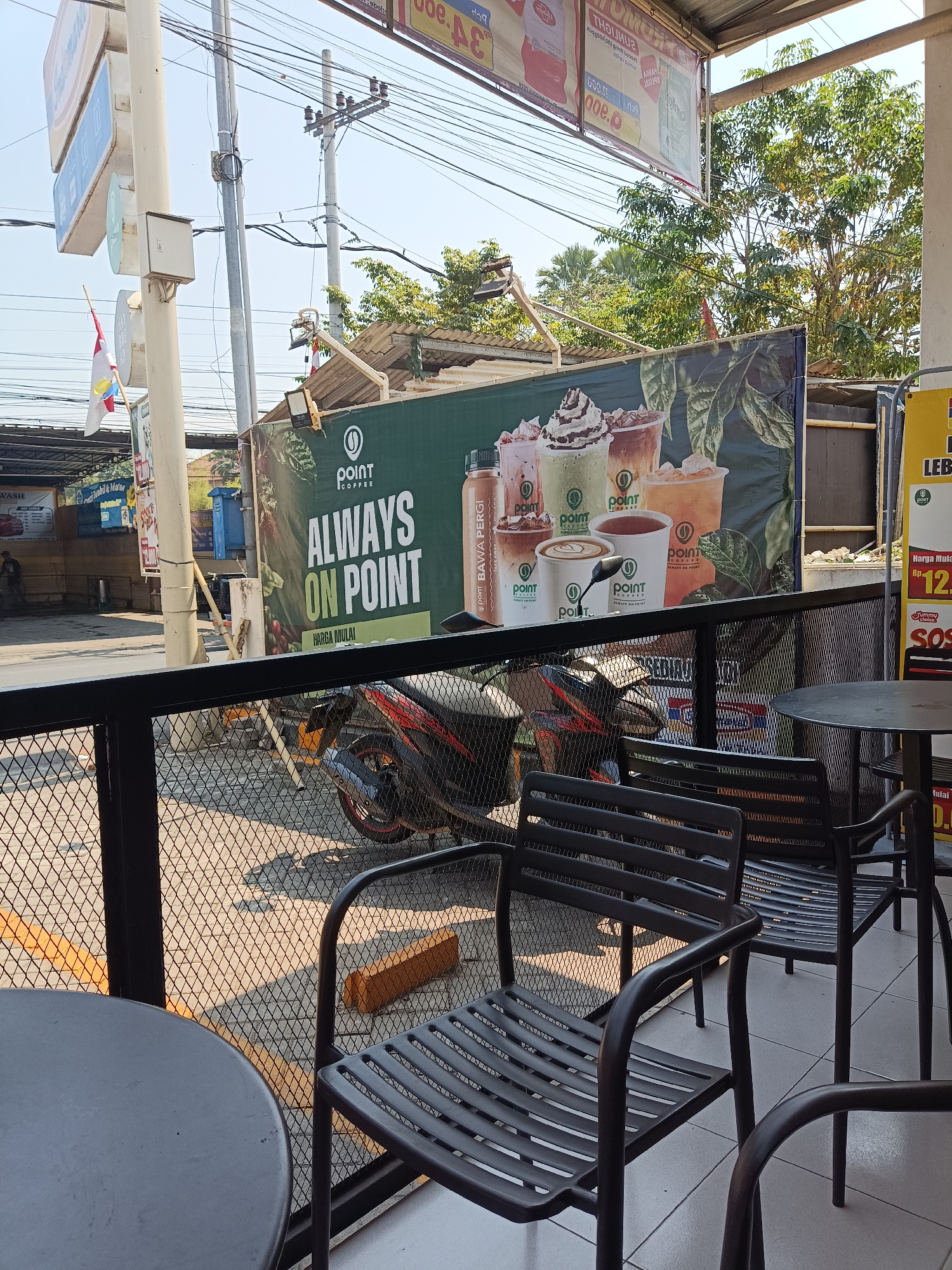 The image depicts an outdoor seating area of a coffee shop, likely located in Jember Kidul, Indonesia, given the contextual hints. In the foreground, several dark metal chairs and a round table are visible, set on a tiled floor. Beyond this seating area, a black metal fence separates it from a paved outdoor space where a red and black motorcycle is parked.
In the background, a large green banner advertising "POINT COFFEE" with the slogan "ALWAYS ON POINT" is prominently displayed. The banner features images of various coffee drinks. To the left of the banner, a white pole with signage and a small flag attached is visible, along with utility poles and overhead electrical wires. Further in the background, trees and parts of buildings can be seen, suggesting an urban or semi-urban environment. The lighting suggests it is daytime, with sunlight casting shadows. There are no discernible people actively engaged in the scene within the frame.