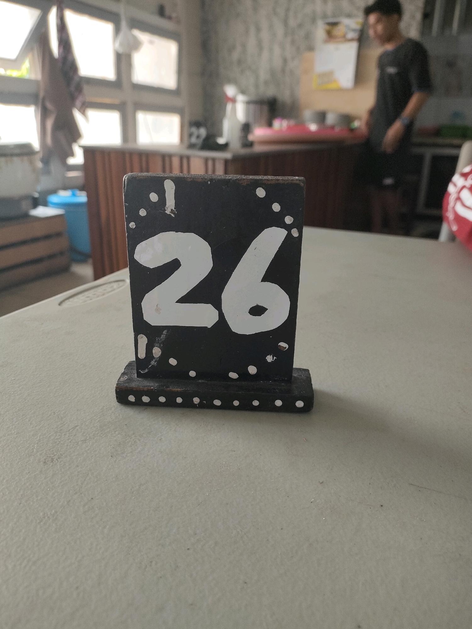 A table number sign, displaying the number "26" in white on a black background, stands on a light-colored, textured surface. The sign has a wooden base and is decorated with small white dots along its edges. In the background, a person in a dark t-shirt and shorts is visible, standing near a counter in what appears to be a casual dining establishment or cafe. The environment is somewhat cluttered, with windows, a blue trash can, and kitchen equipment visible in the blurred background, suggesting an indoor setting.