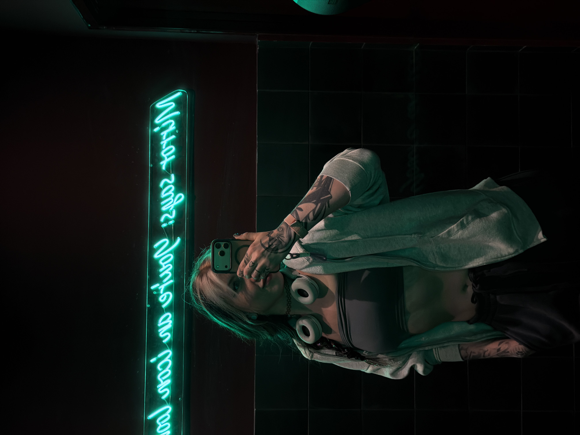A photograph taken in an interior setting, likely in the Čukarica Urban Municipality of Belgrade, Serbia. The scene features a woman taking a selfie. She is wearing headphones around her neck, a sports bra, and a hoodie. Tattoos are visible on her arms. She is holding a smartphone up to take the photo. Next to her is a vertical neon sign with text in a handwritten style. The background consists of a dark wall and tiled surfaces.