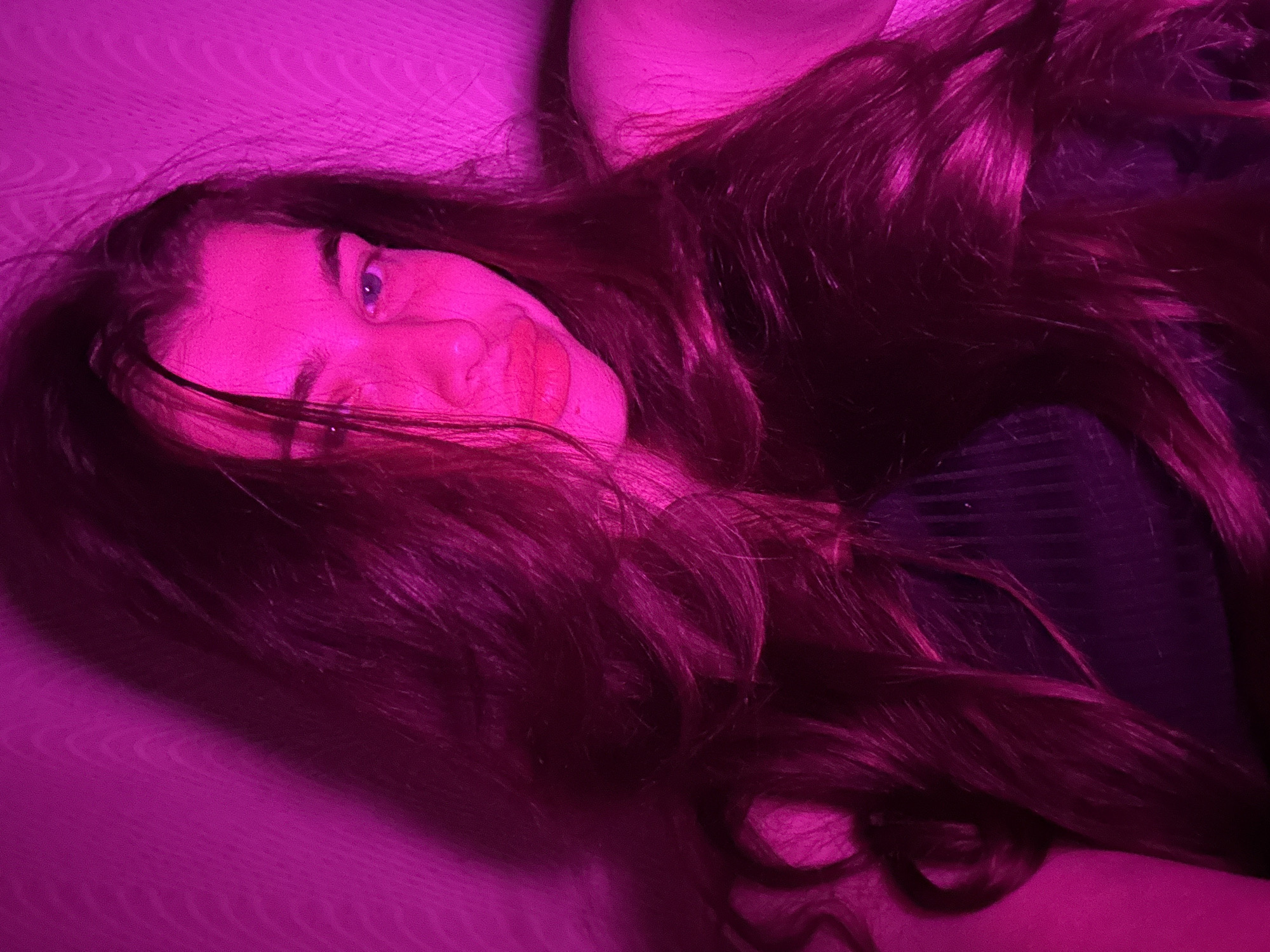 A young woman with long, dark, wavy hair is pictured from the chest up. She is looking towards the camera with her head tilted slightly to the right. Her skin is illuminated by a strong pink light, which also casts shadows on the wall behind her. She is wearing a dark, possibly black, top with a ribbed texture. Her left eye is more visible than her right, and her lips are slightly parted. The background appears to be a textured wall. There are no discernible objects or other people in the frame. The scene is devoid of any specific geographical markers that would indicate its location in Мещёры, Russia.