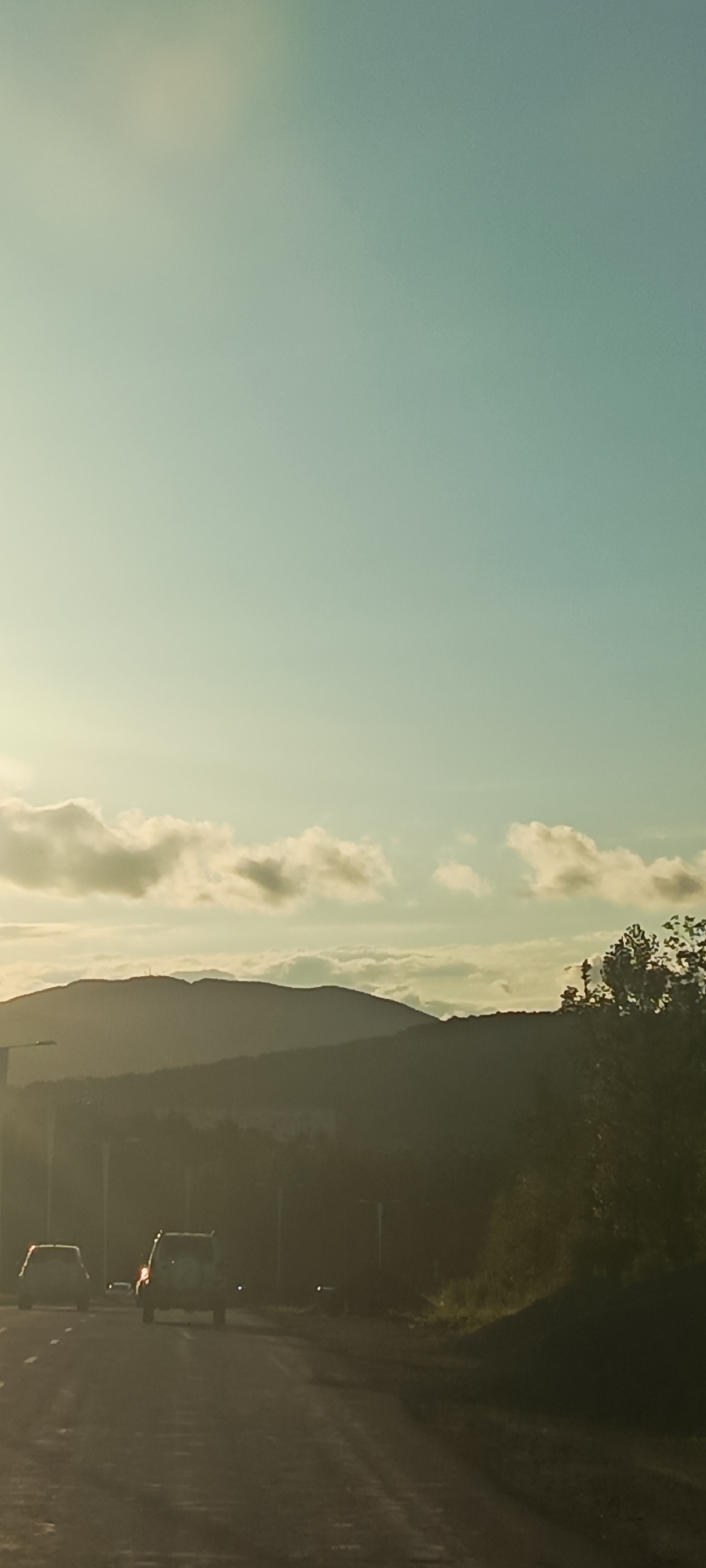 The image depicts a serene sunset over a mountainous landscape, likely on Russky Island, Russia. The sky is a gradient of pale yellow near the horizon to a soft teal overhead, with scattered, wispy clouds catching the golden light. In the foreground, a dimly lit road stretches forward, with two vehicles visible. The vehicle in the foreground is a white SUV with a spare tire mounted on the back, and a second white vehicle is visible further ahead. The road appears to be asphalt, with some reflections from the low light. In the middle ground, silhouetted trees and foliage form a dark line against the illuminated sky and mountains. The mountains themselves are layered, with the furthest peaks appearing as soft, hazy outlines. The overall impression is one of tranquility and natural beauty as day transitions into evening.