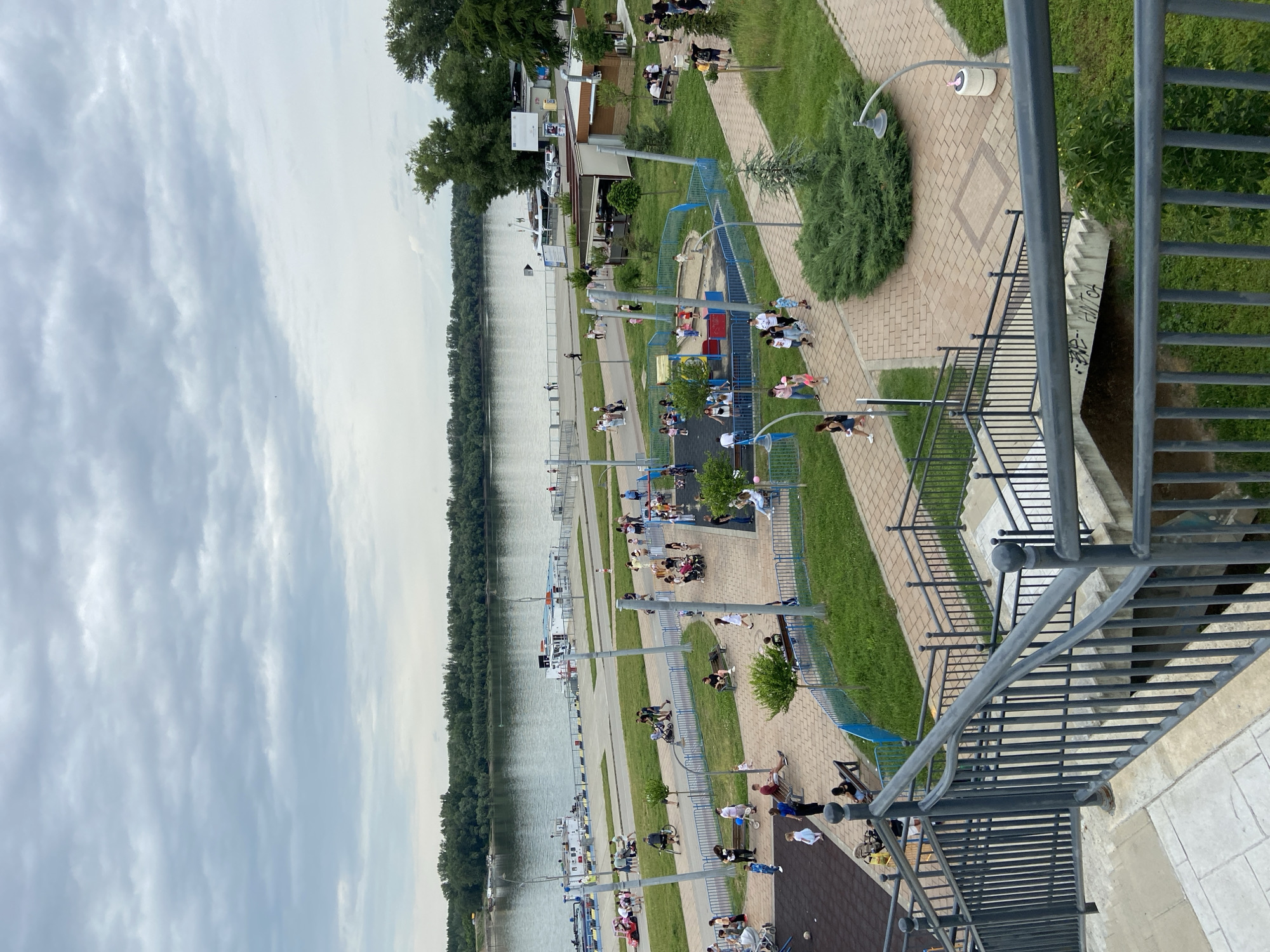 The image shows an elevated view of the Danube Riverbank in Ruse, Bulgaria. The Danube River is visible on the left side of the image, lined with trees on its banks. Along the riverbank, there's a paved promenade area with benches, playgrounds, and recreational facilities. People are scattered along the promenade, some sitting on benches, others walking or gathered in small groups near the playground equipment. The lighting suggests it is daytime. The image is taken from a high vantage point, possibly from a building or structure overlooking the riverbank. Metal railings and stairs are visible in the foreground, indicating the viewpoint's position.