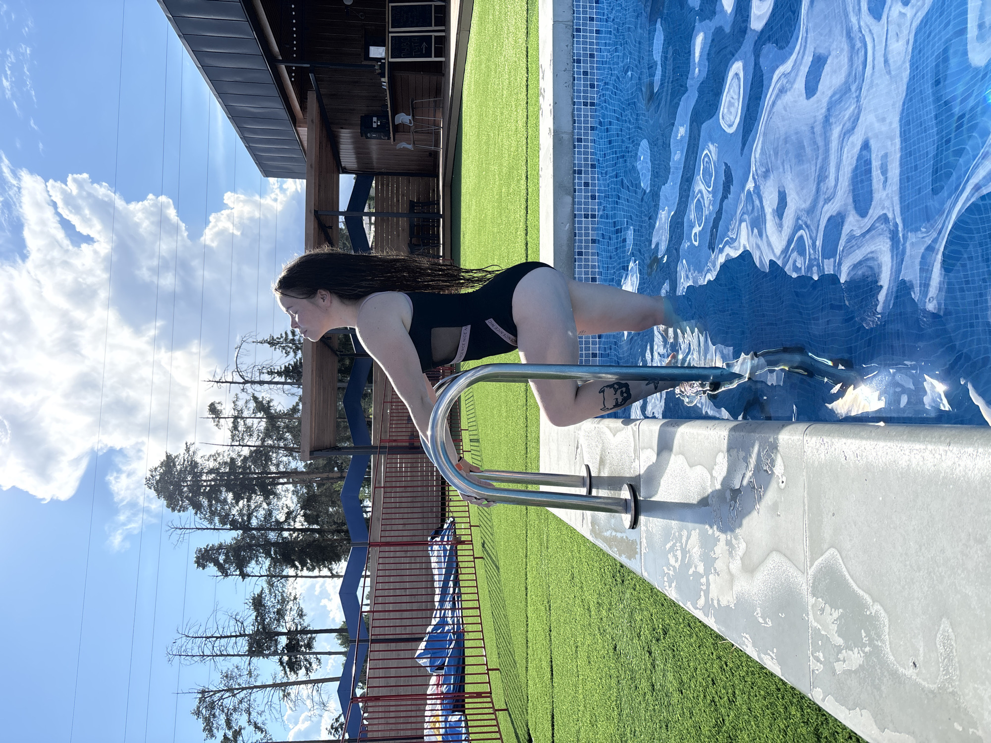 A woman in a black swimsuit is climbing out of a swimming pool, using a metal ladder. The pool has blue tiles and rippling water. Artificial turf surrounds the pool. Beyond the pool area is a red fence, trees, and a building with a dark roof. The sky is blue with scattered clouds. The photograph was taken during daylight. The location is Votkinsk, Russia.