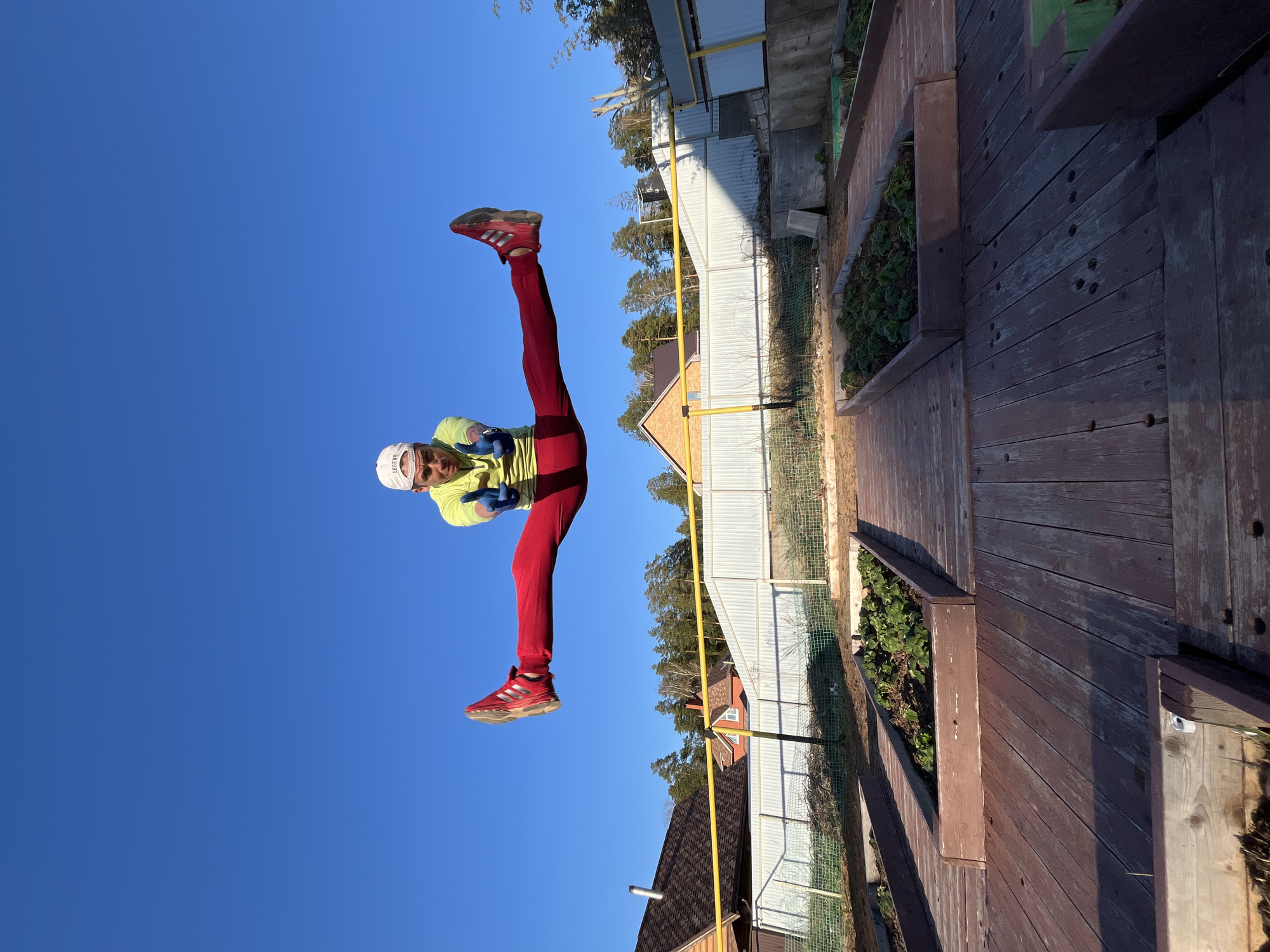 A man, wearing red pants, red shoes, a yellow long-sleeved shirt, blue gloves, and a white cap, is doing the splits in front of a yellow gas pipe running along a fence, presumably in Chaikovsky, Russia. He is positioned against a bright blue sky. The backdrop also includes parts of houses, trees, and a wooden structure with plant boxes.