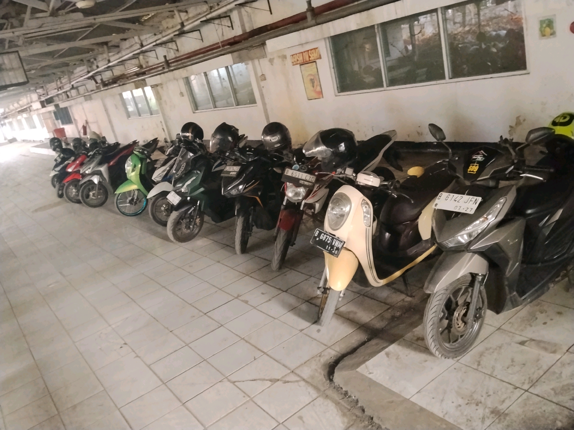 The image shows a row of parked motorcycles in what appears to be an indoor parking area. The motorcycles are lined up against a wall with windows. The floor is tiled and appears to be somewhat dirty. Some of the motorcycles have helmets on them. There are signs on the wall. The lighting is artificial and somewhat dim, suggesting an indoor or covered space, possibly a parking garage or a covered outdoor area. The overall impression is one of an organized, if somewhat utilitarian, parking facility. The image does not provide any specific visual cues to confirm the location as Suka Nagara, Indonesia, but the presence of Indonesian license plates on some of the motorcycles suggests a Southeast Asian context.