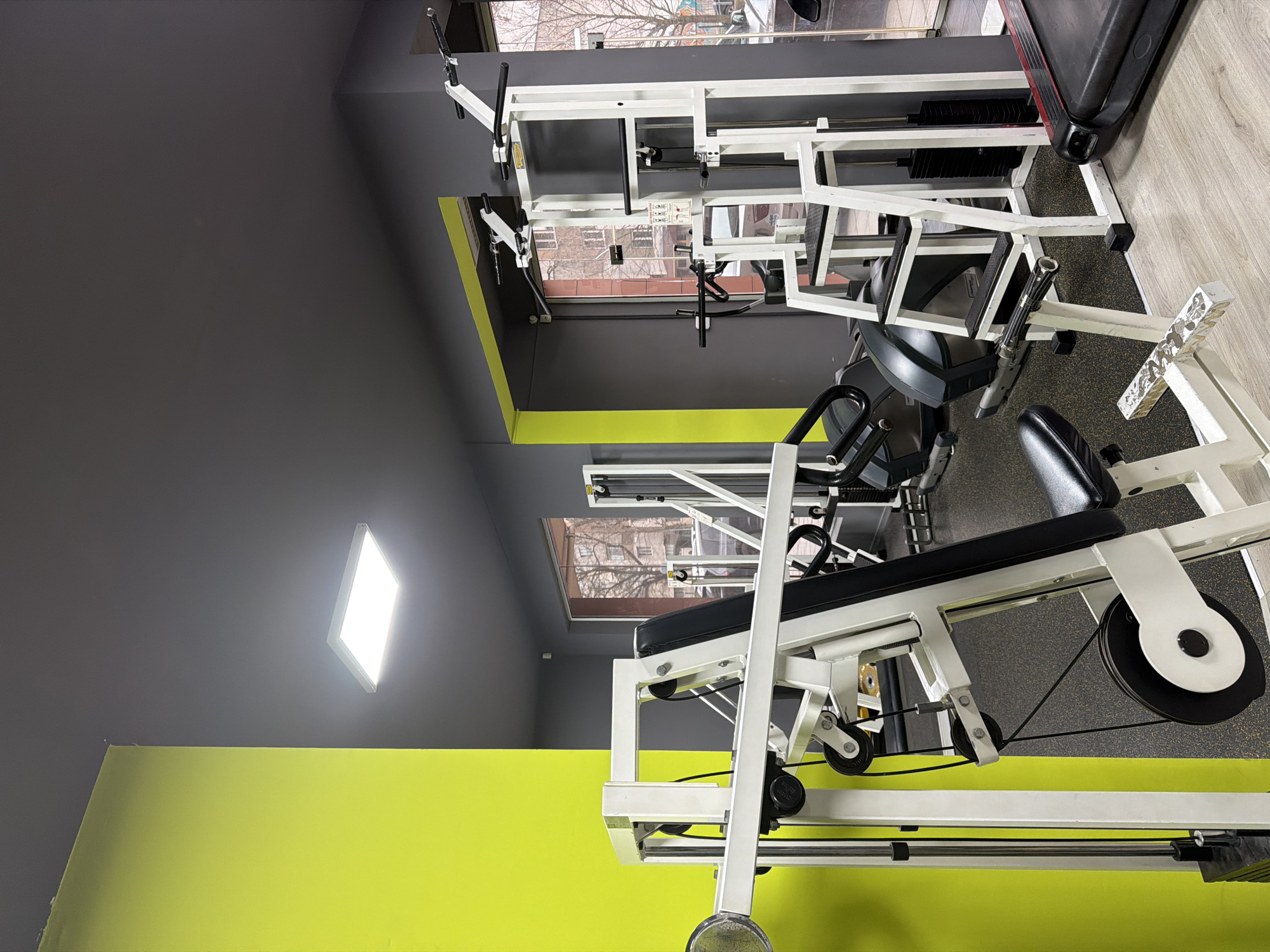 The image shows a gym in Sofia, Bulgaria, with several white weight machines. The walls are two-toned, with gray above a yellow-green section. A rectangular ceiling light illuminates the room. Through the windows, bare tree branches and buildings can be seen. The flooring appears to be wood-look planks and rubber flooring with a speckled pattern.