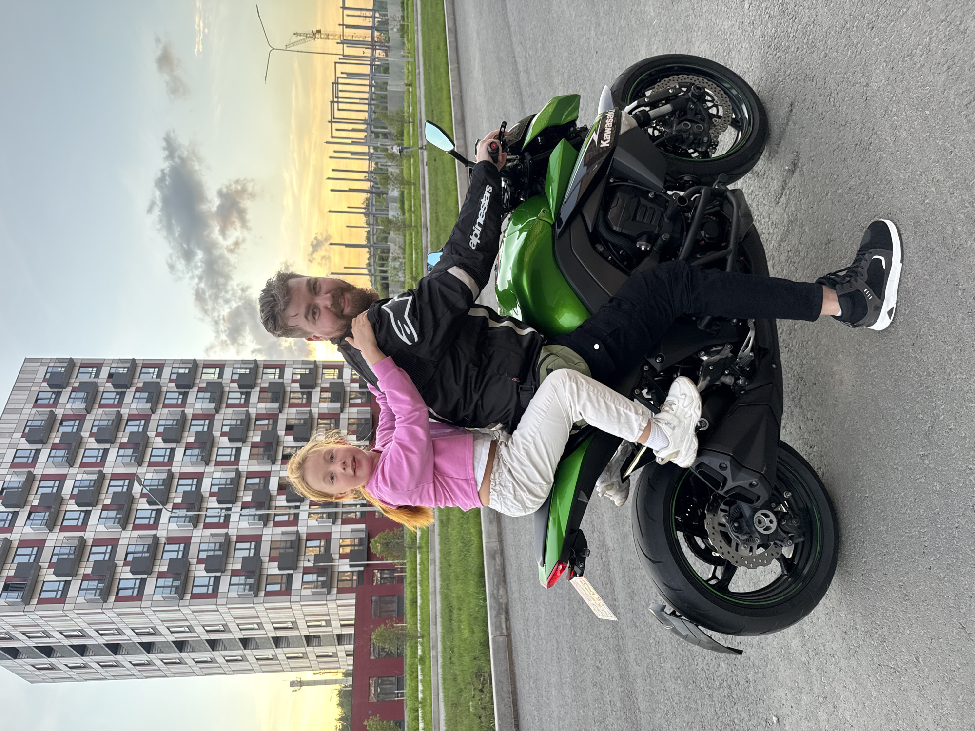 The image shows a man and a young girl on a green and black motorcycle. The man is in the front, wearing a black motorcycle jacket with "Alpinestars" and "Kawasaki" visible. He has a beard and is smiling at the camera. The girl is sitting in front of him, facing the camera, wearing a pink hoodie and white pants. She has blonde hair and appears to be smiling.
They are positioned on a paved surface, likely a road or parking lot, with a modern, multi-story building on the left side of the frame. The building has a repetitive pattern of windows and balconies. In the background, there are construction elements, possibly scaffolding or unfinished structures, and some trees.
The time of day appears to be late afternoon or early evening, with a bright, hazy sky suggesting sunset or sunrise. The lighting is warm, casting a soft glow on the scene. The overall mood seems happy and relaxed, with the man and girl enjoying their time together on the motorcycle. The location is specified as Veliky Novgorod, Russia, which aligns with the urban and suburban elements visible in the background.