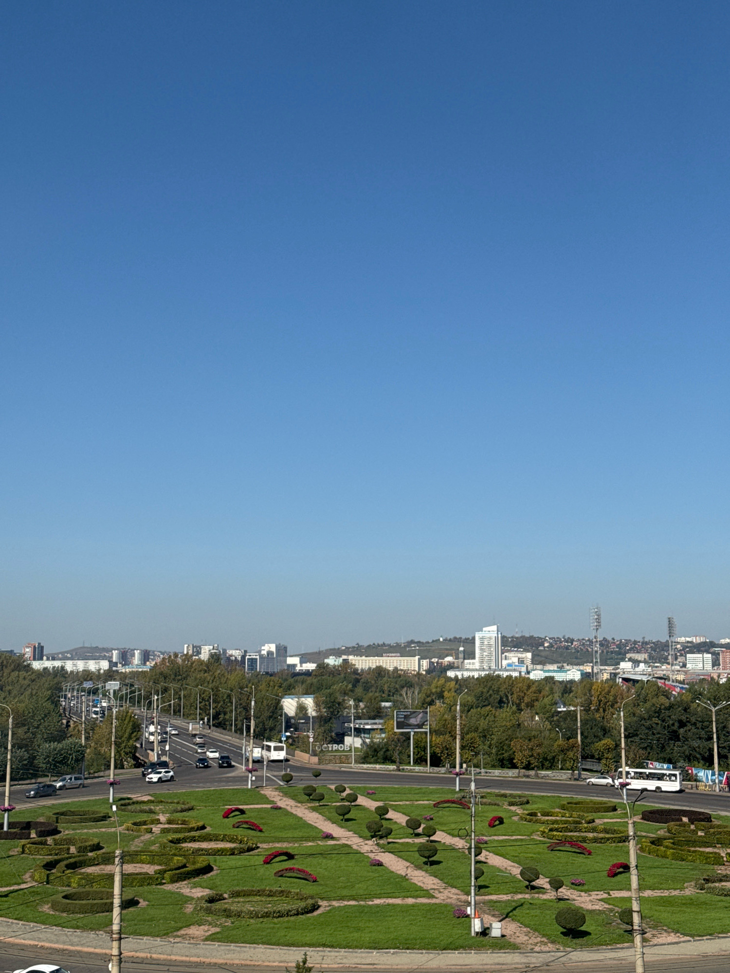 The image depicts a panoramic view of the city of Krasnoyarsk, Russia, on a clear, sunny day. In the foreground, a meticulously maintained park area with green lawns, manicured hedges, and decorative flower beds is visible. Several pathways wind through the park, and lampposts are strategically placed. Beyond the park, a multi-lane road carries vehicular traffic, including cars and buses. A bridge spans across the road in the middle ground, with more trees and foliage on either side. In the background, the cityscape of Krasnoyarsk unfolds, featuring a mix of modern high-rise buildings, residential complexes, and industrial structures. The urban landscape is surrounded by rolling hills covered in dense forest. The sky is a clear, vibrant blue.