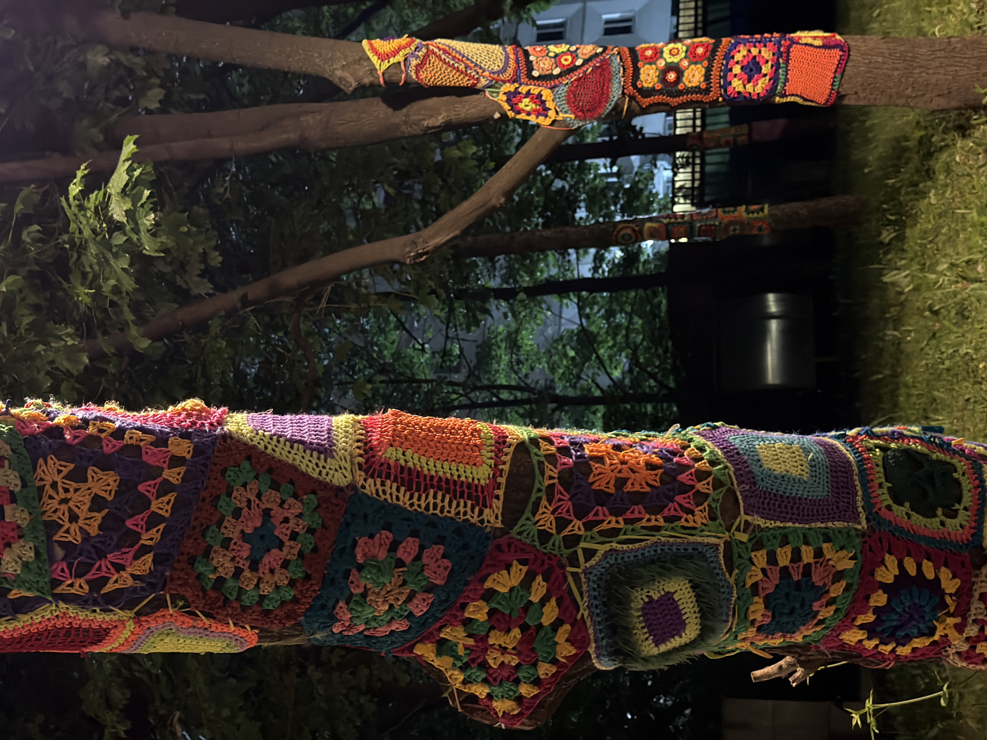 The image shows tree trunks wrapped in colorful knitted squares, likely yarn bombing, in an outdoor setting. The location is in Moscow, Russia, judging by the architecture in the background. The scene is at night with artificial lighting. There is green vegetation visible, including tree leaves and grass. A metallic object is partially visible between the trunks. No people are visible in the image.