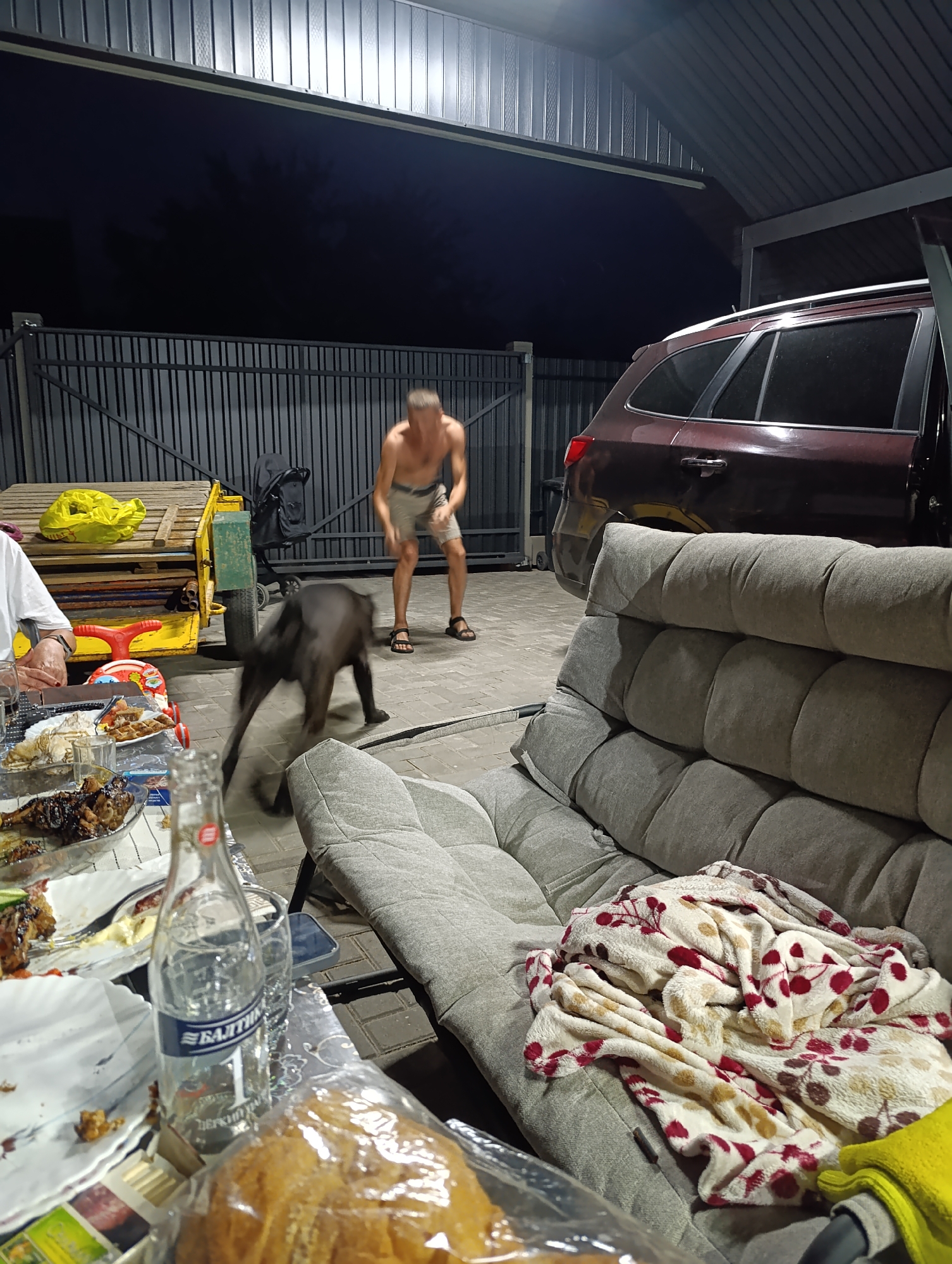 An evening scene in Slutsk, Belarus, takes place on a paved surface under a metal-roofed overhang. In the background, a man in shorts is silhouetted against a dark sky, standing near a fence. In the midground, a dark-colored SUV is parked. A plush gray outdoor sofa occupies the right side of the frame, draped with a patterned blanket. In the foreground, a table is laden with food, plates, and a bottle of water. A black dog is captured mid-motion, appearing to be playing or interacting with someone out of frame. A yellow push toy and a large yellow bag are visible on a wheeled cart. The overall atmosphere suggests a casual outdoor gathering.